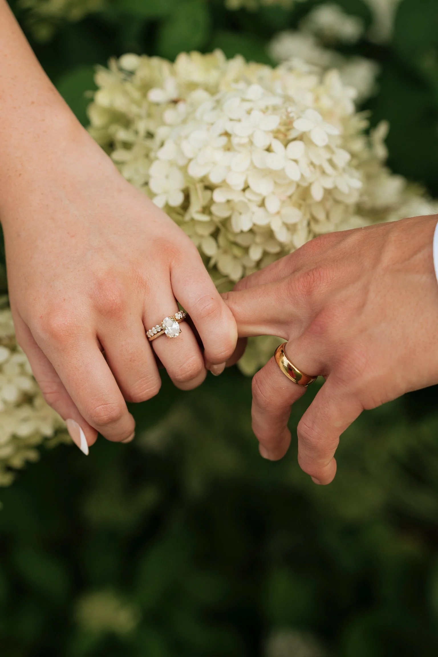 Close-up of a bride and groom holding hands outdoors, showing wedding rings amid green foliage and white flowers in the background.