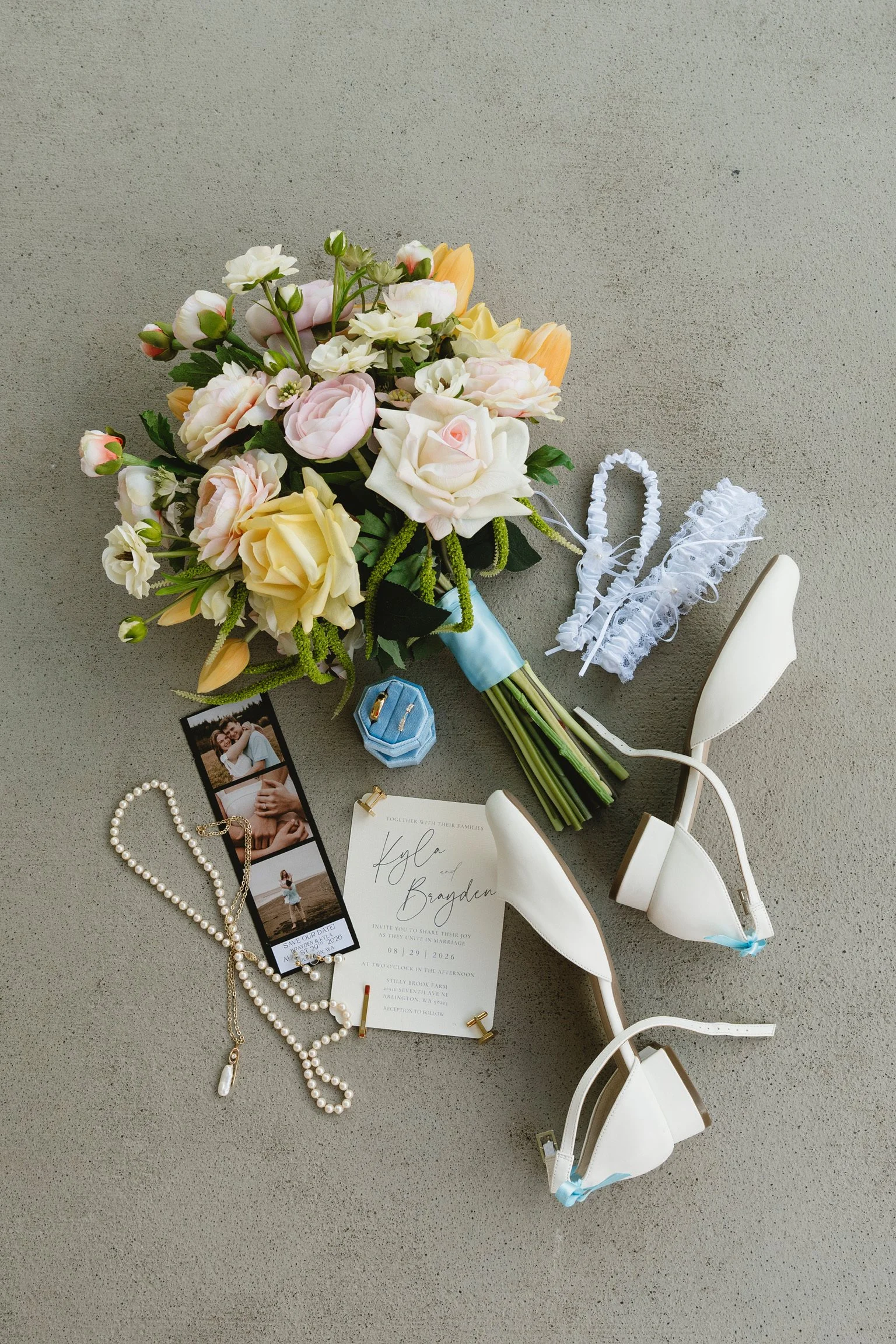 Wedding invitation, bouquet of roses, jewelry, earrings, garter, and white high-heeled shoes on a grey surface.