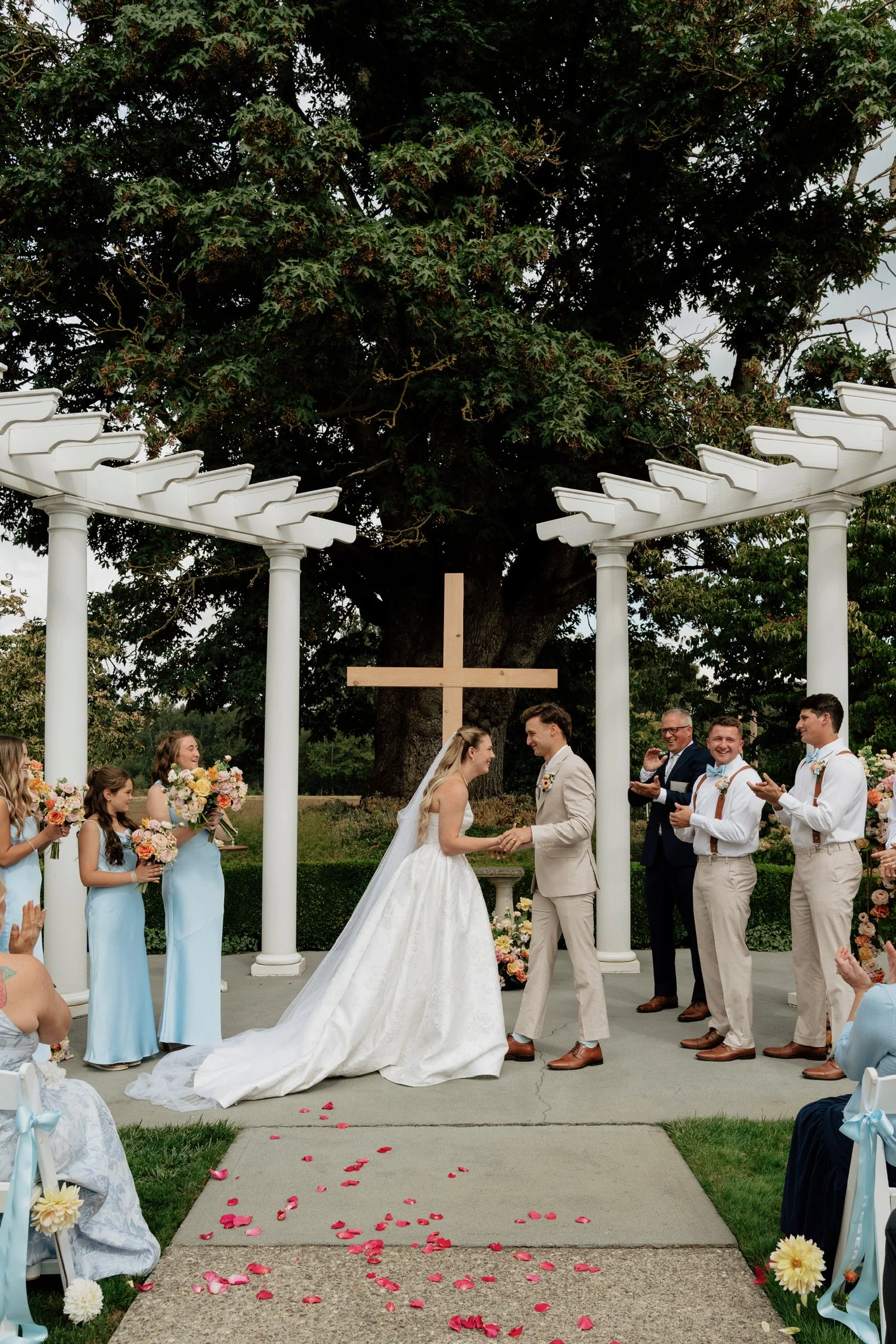 A wedding ceremony taking place outdoors under a large tree with a white pergola overhead. The bride and groom are holding hands in front of officiant. Bridesmaids in blue dresses hold bouquets and stand on the left, groomsmen in beige pants and whit