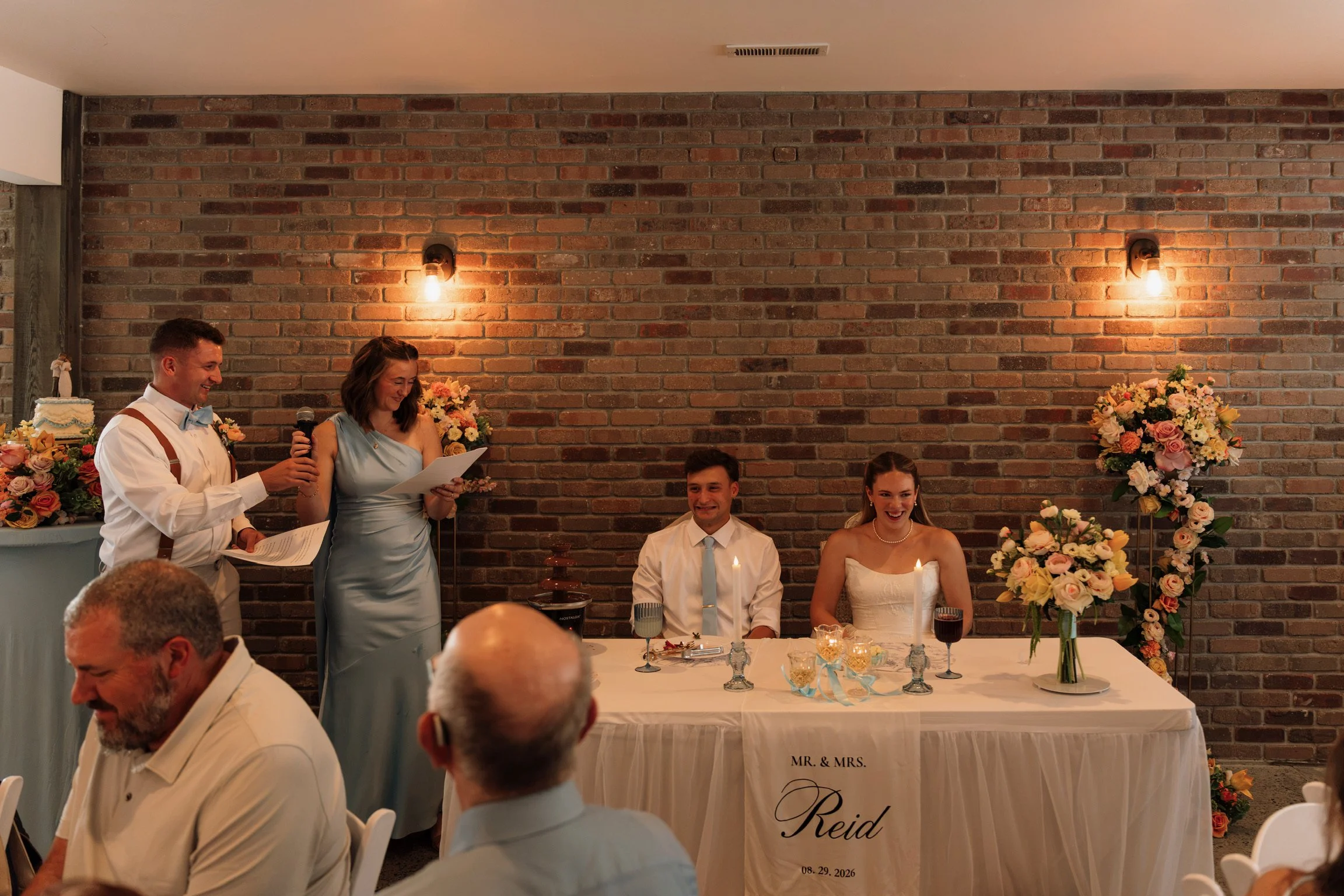 A wedding reception scene with a bride and groom sitting at a table with candles and floral arrangements. A woman in a light blue dress is reading from a paper while a man in suspenders and a bow tie smiles next to her. The wedding date is August 29,