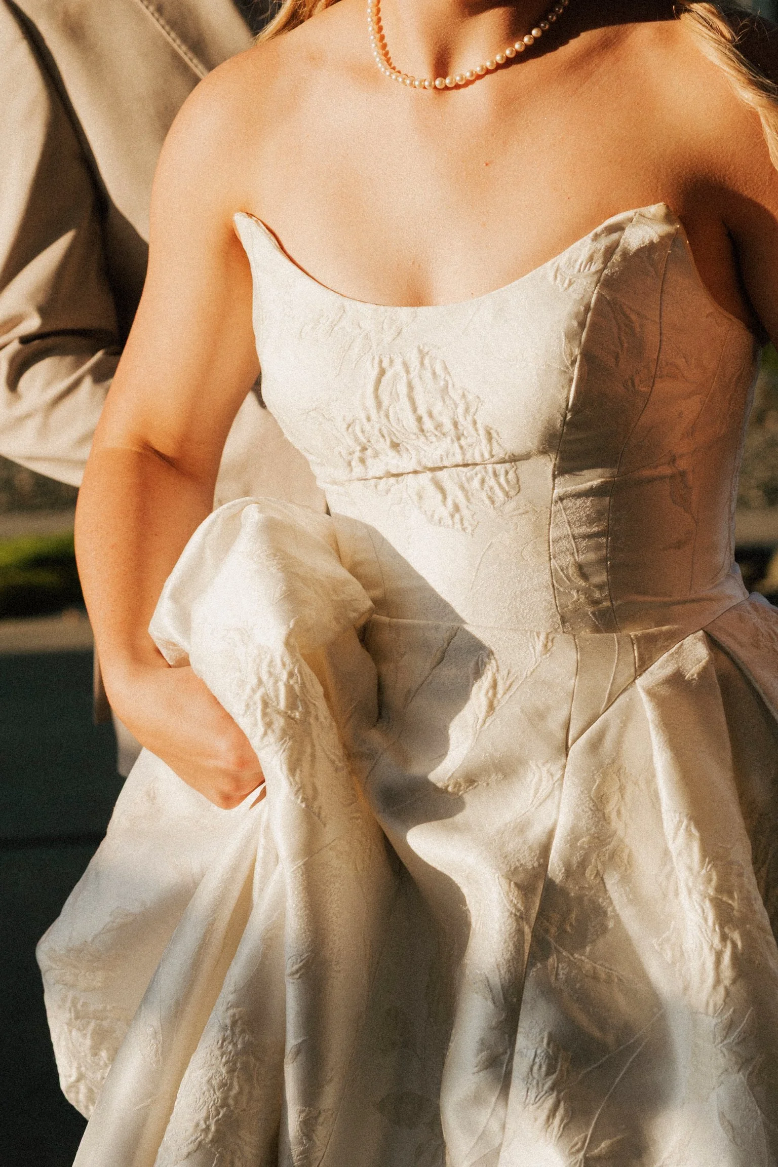Close-up of a woman in a cream-colored, textured gown with a fitted bodice and puffed sleeves. She is wearing a pearl necklace and has light brown hair. The background is blurred, with a person in a beige suit partially visible.