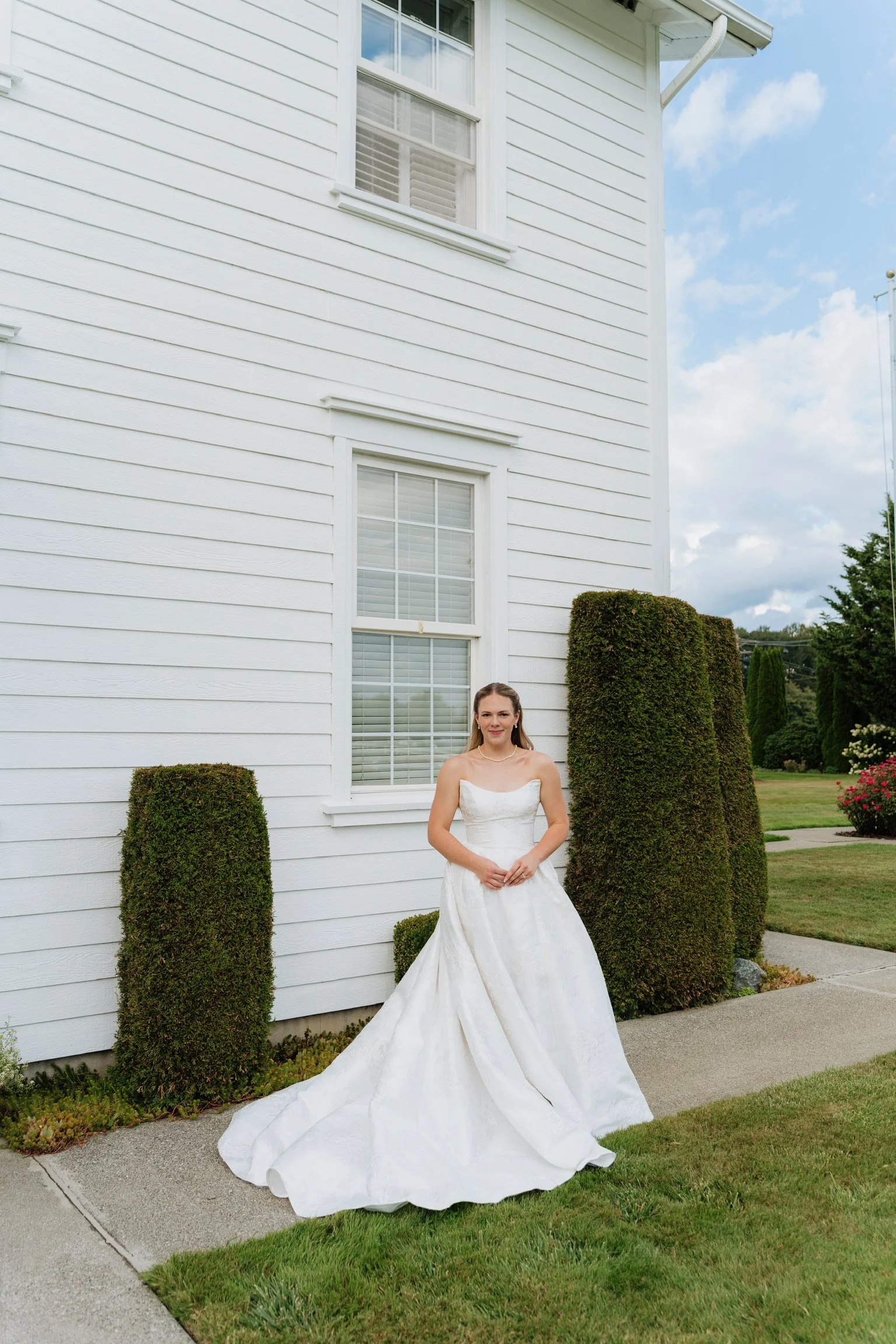 A woman in a white wedding dress standing outside near a white house with double-hung windows and well-manicured bushes, under a partly cloudy sky.