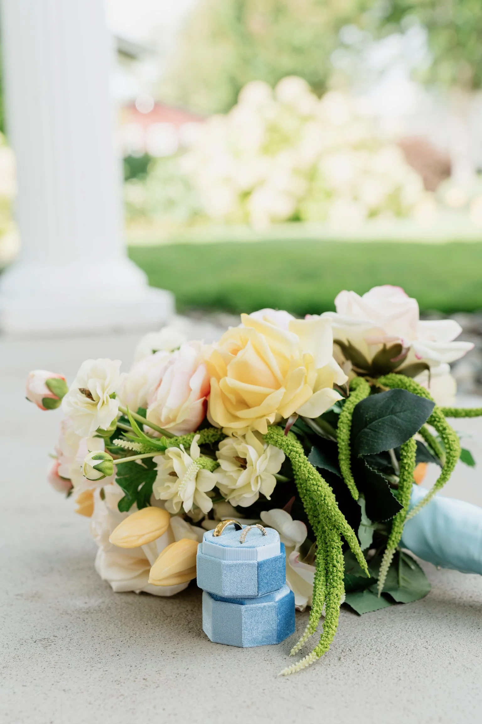 A bouquet of white and cream flowers, including roses and tulips, with green accents, placed on a gray surface near a window with a blurred outdoor background; in front of the bouquet are small velvet ring boxes, one blue and one pale blue, containing wedding rings.