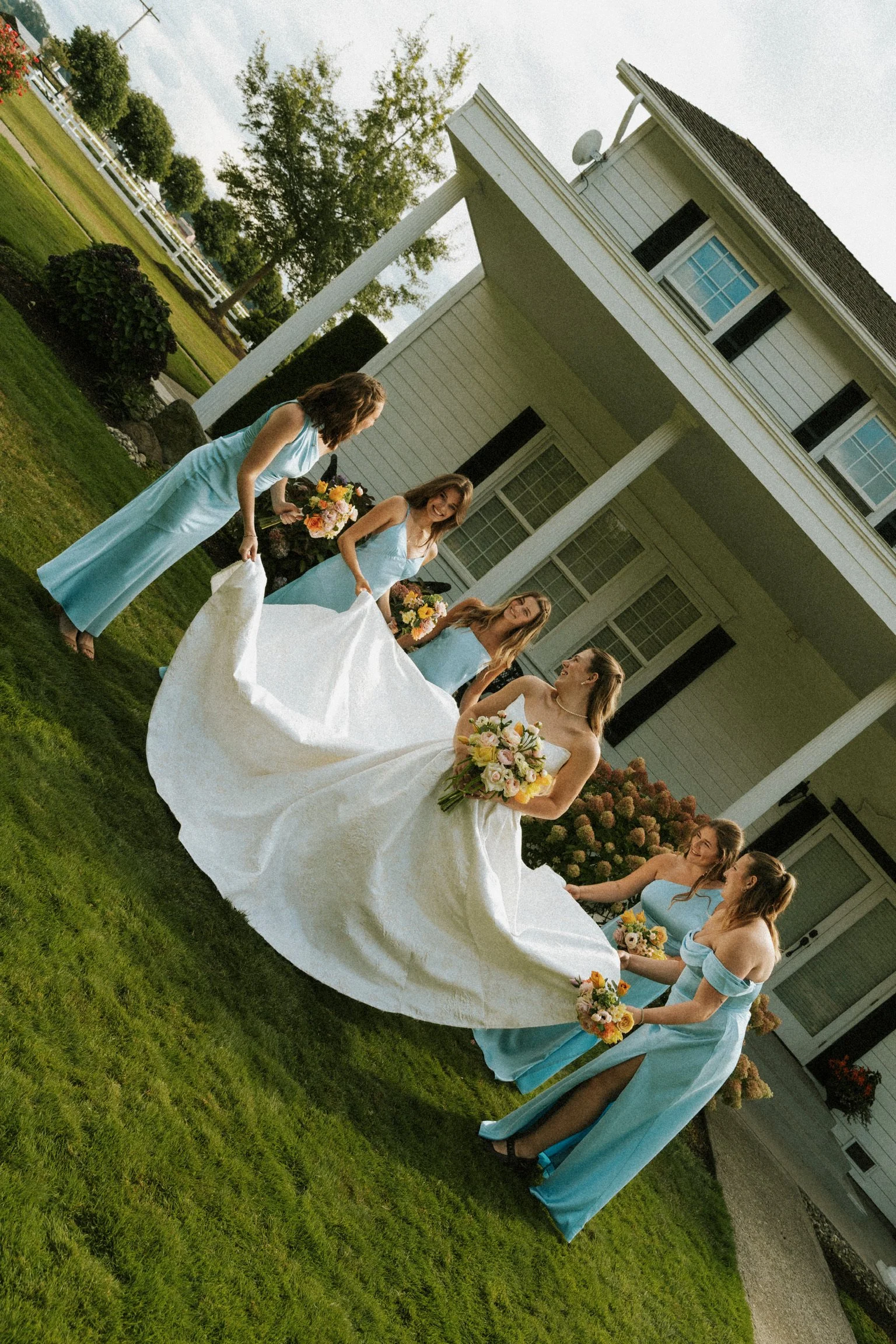 A group of women in light blue dresses and a bride in a white wedding gown standing on a well-maintained green lawn in front of a white house. The women are holding bouquets of flowers and appear to be participating in a wedding celebration.