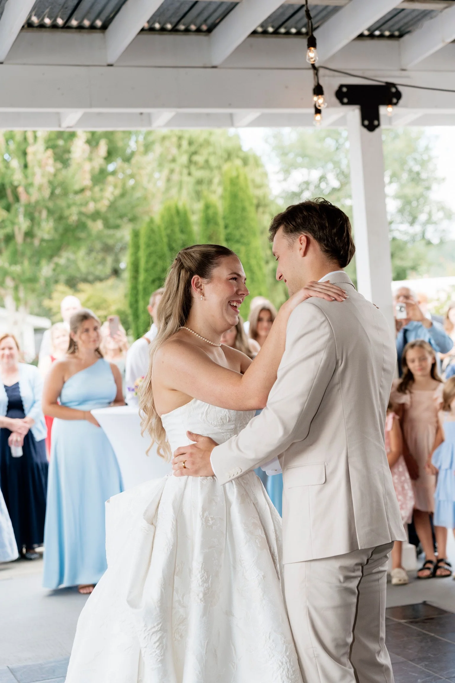 A bride and groom sharing a dance at their wedding reception, surrounded by guests outdoors under a white pavilion.