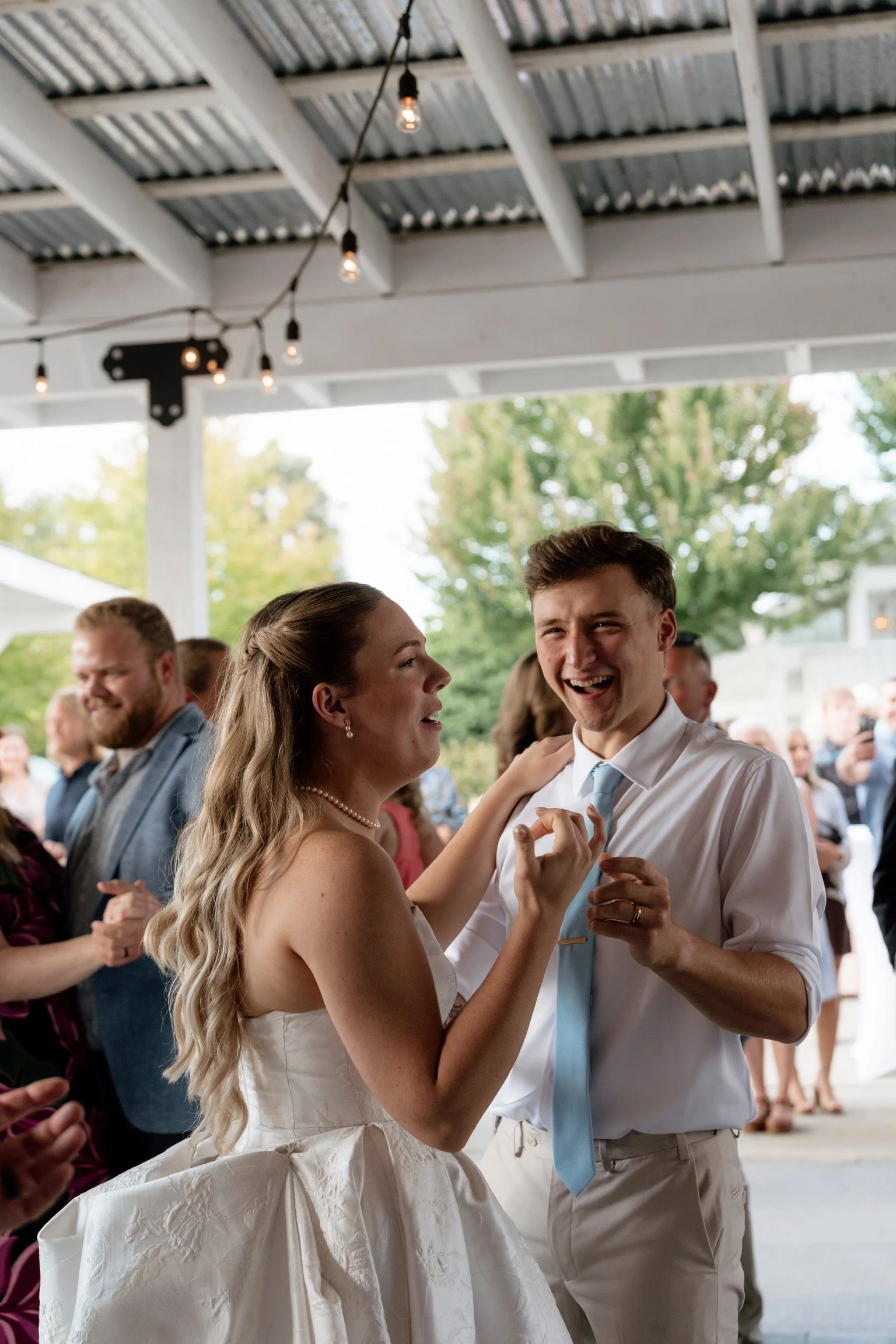 A bride and groom happily dancing at their wedding reception, with guests in the background enjoying the celebration outdoors under a corrugated metal roof, decorated with string lights.