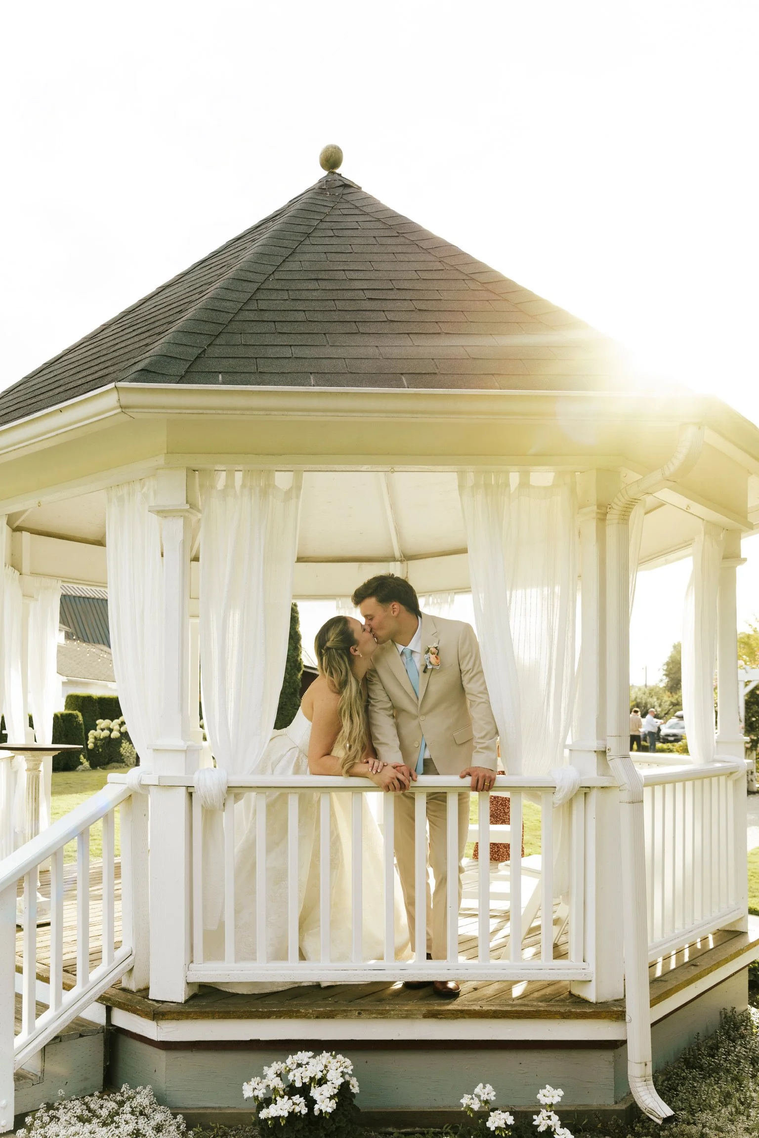 A bride and groom share a kiss on a white gazebo with sheer curtains, outdoors during daytime, with sunlight illuminating the scene.