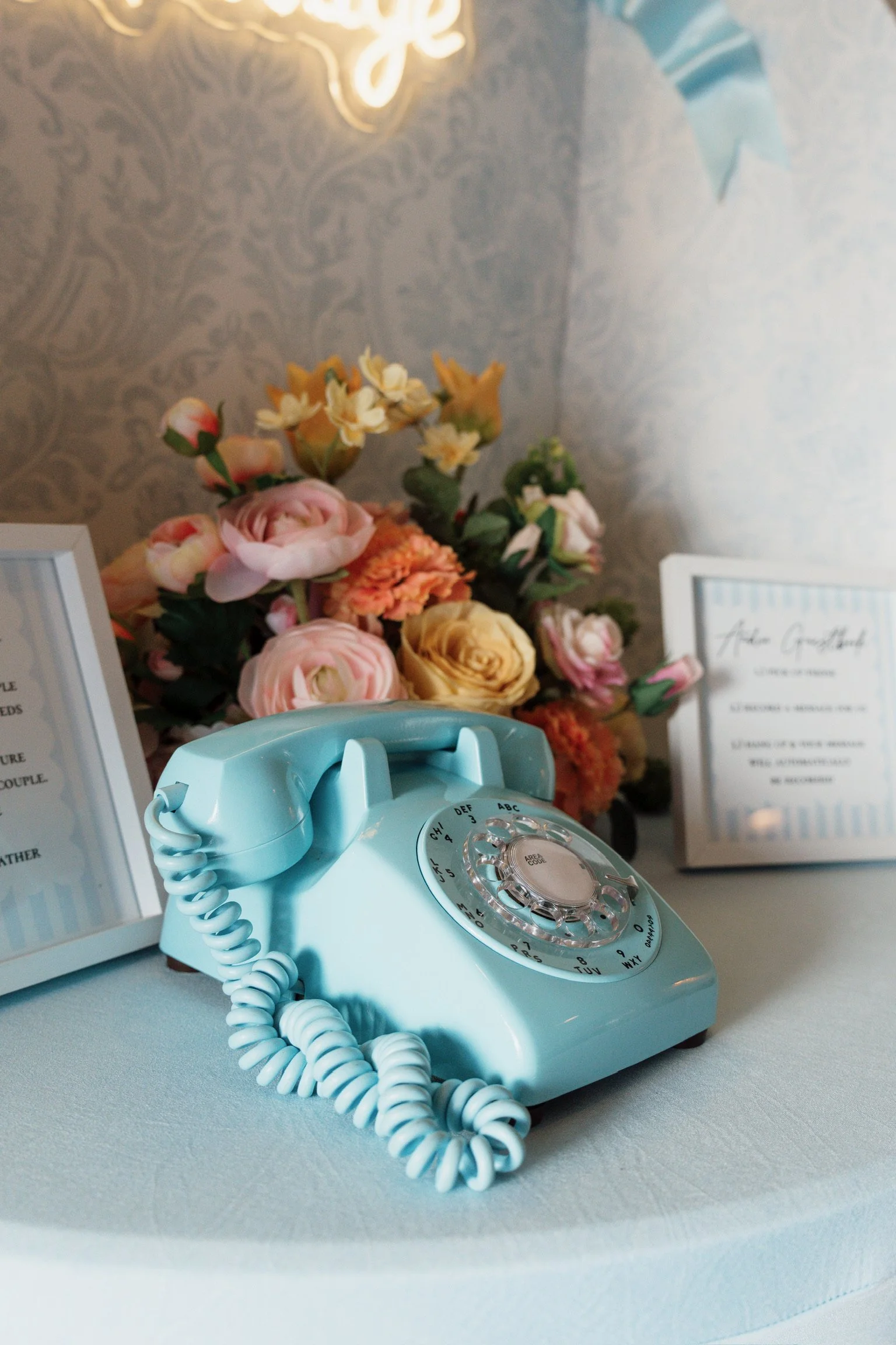 A pastel blue rotary telephone on a white surface with framed signs and a bouquet of pink and yellow flowers in the background.