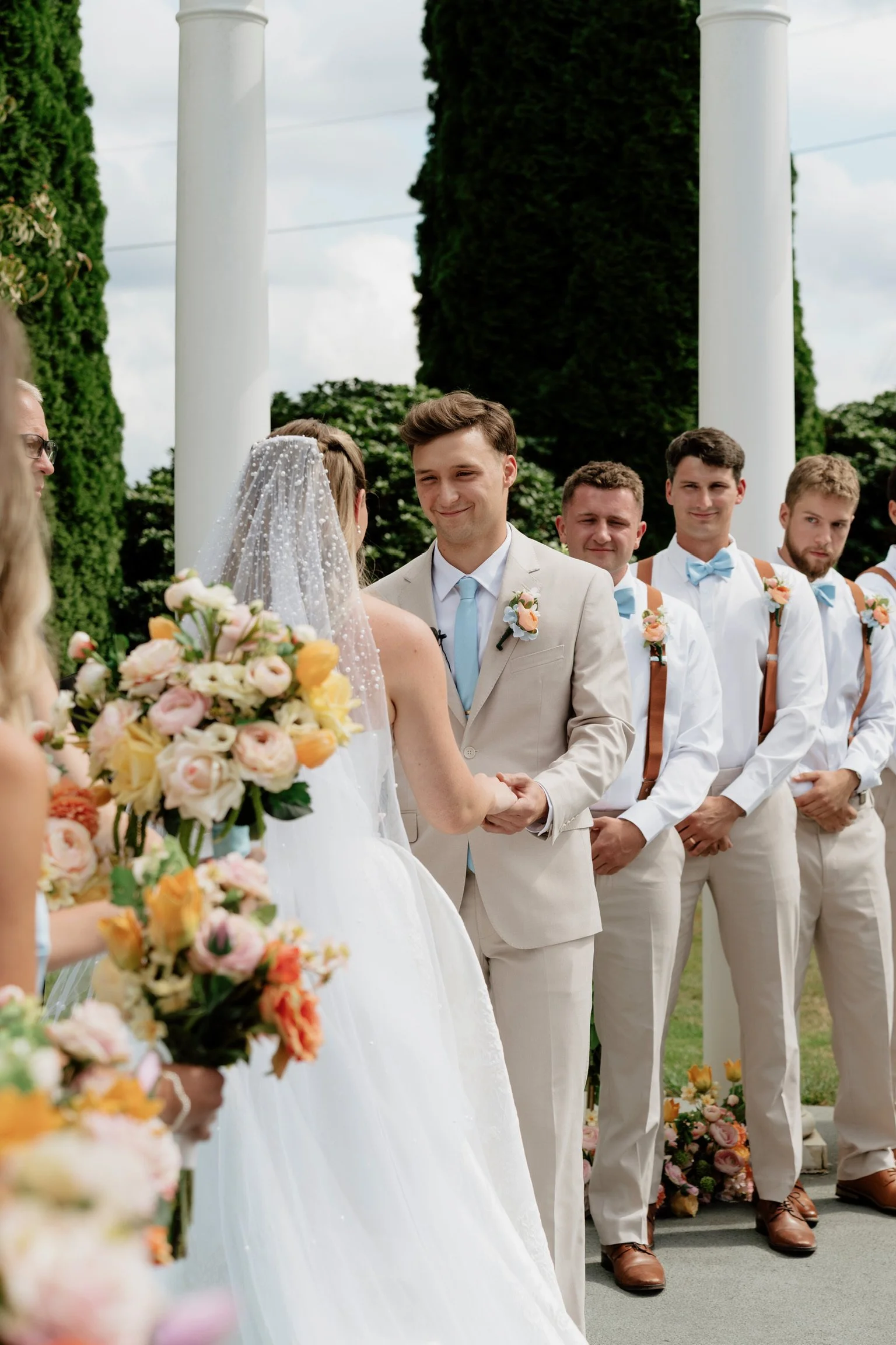 A wedding ceremony outdoors with the bride and groom holding hands, surrounded by bridesmaids and groomsmen. The groom is smiling, wearing a light beige suit with a blue tie and boutonniere. The bride wears a white wedding dress and veil, holding a b