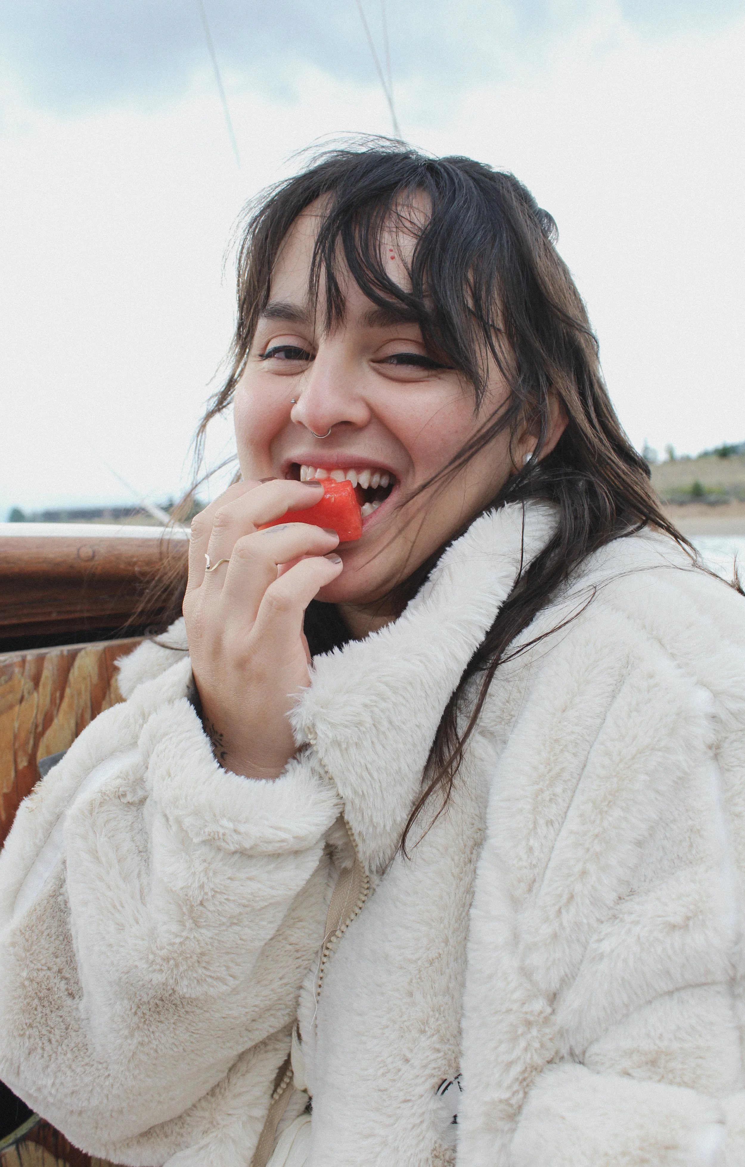 A woman with dark hair, a nose piercing, and a playful smile is biting into a slice of watermelon while wearing a cream-colored fluffy jacket outdoors.