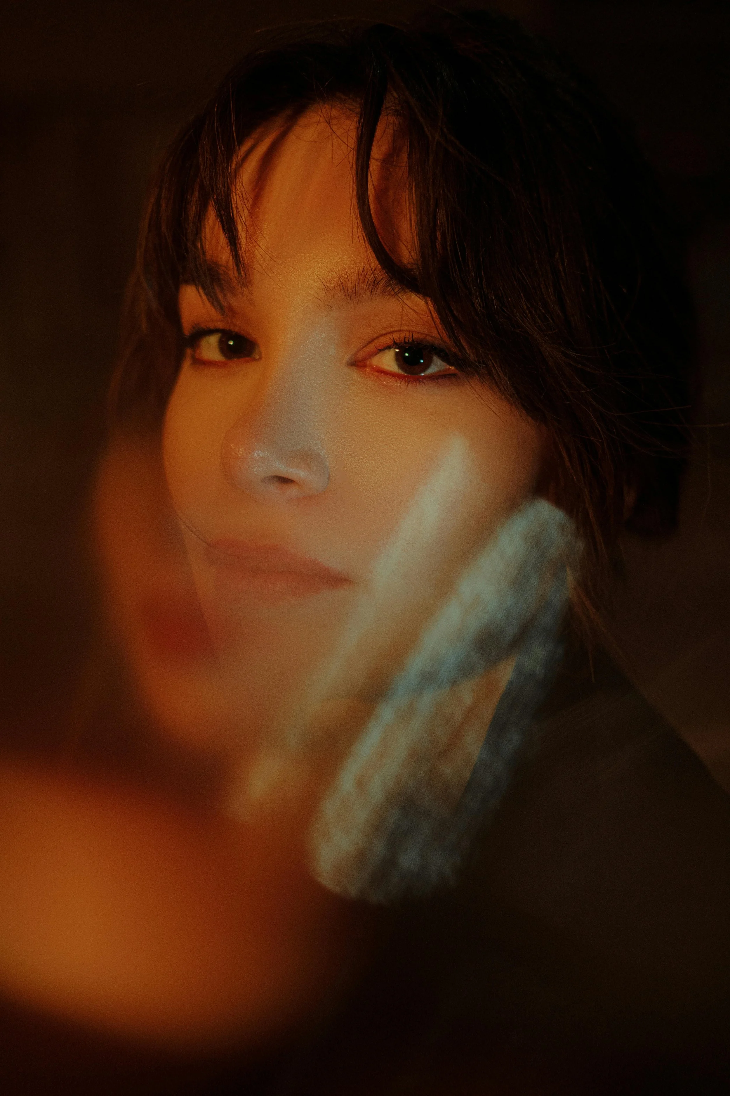 A close-up portrait of a woman with dark hair and light makeup, partially covered by a blurry object in the foreground.