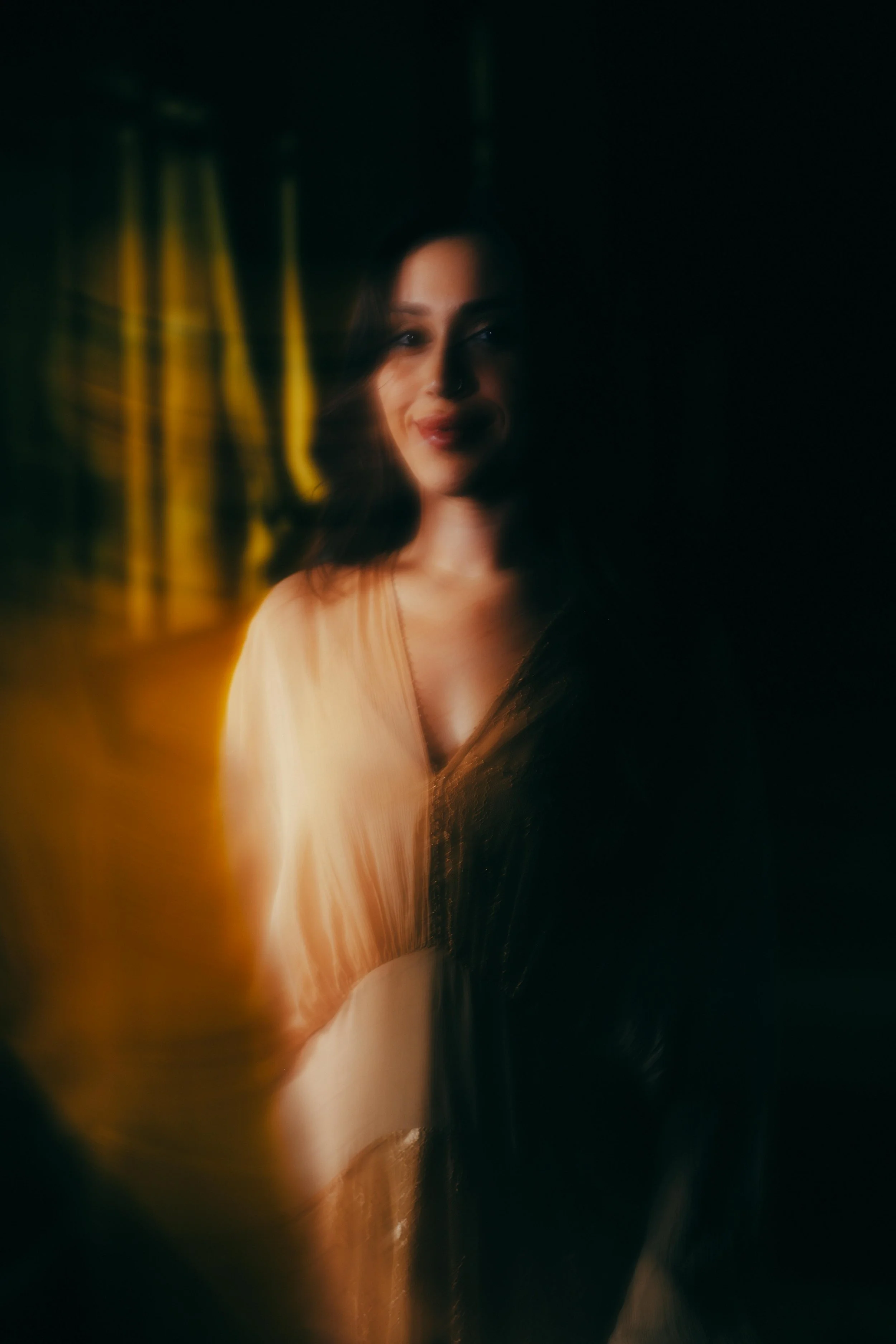 A woman with dark hair smiling in a dimly lit room with reflections on a glass surface.