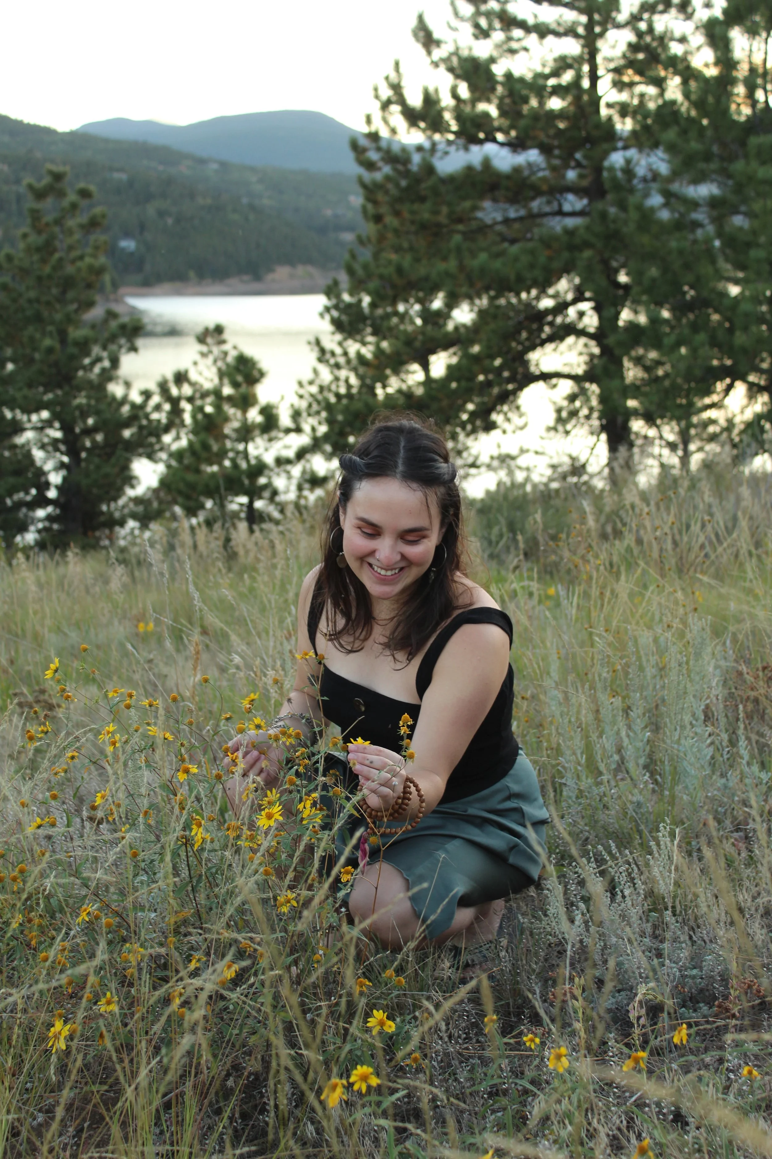 A young woman with dark hair, wearing a black top and teal shorts, kneeling in a field of yellow wildflowers near a lake with pine trees and mountains in the background, during late afternoon or early evening.