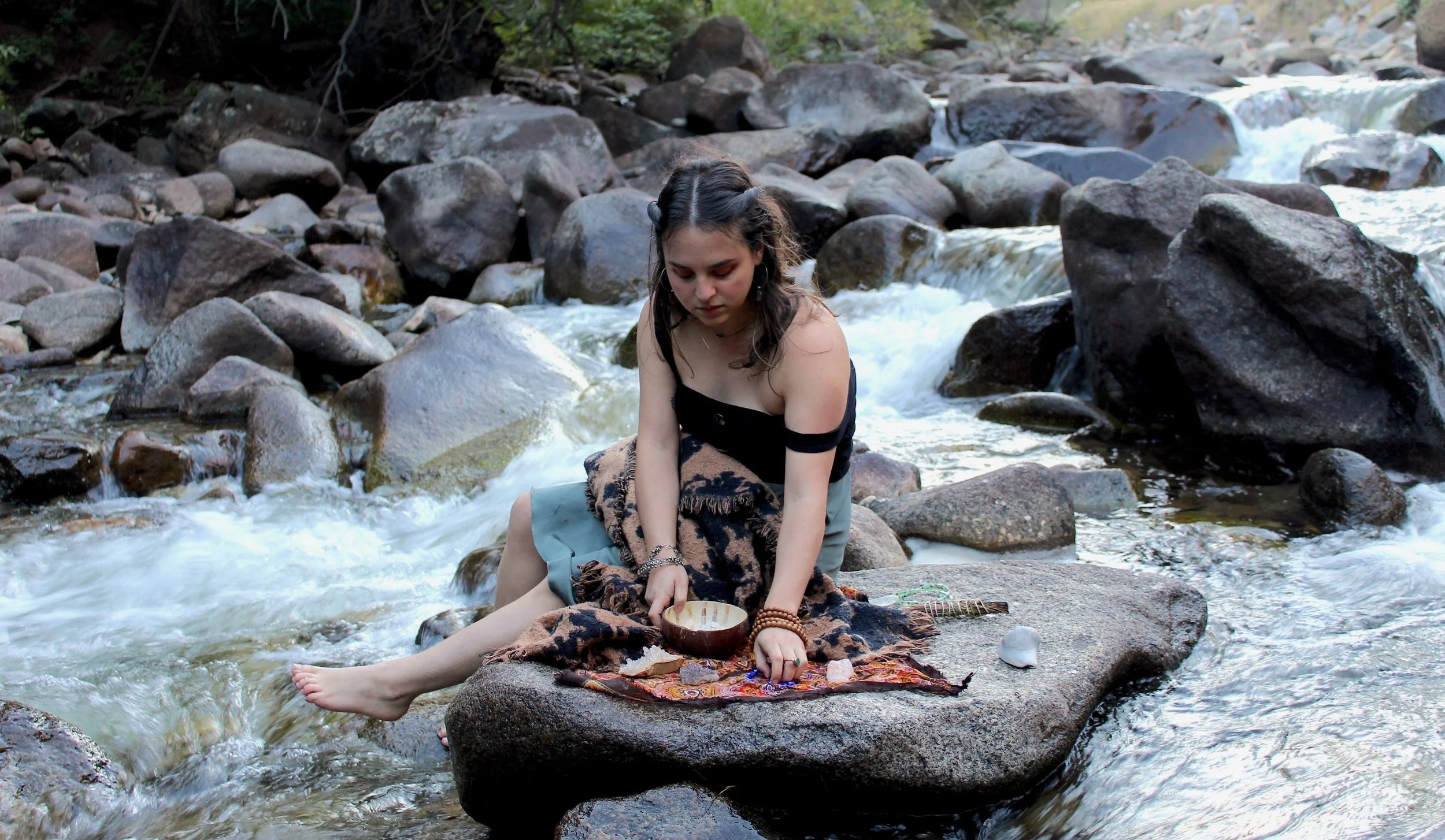 A young woman with dark hair and hoop earrings sitting on a rock in a river, surrounded by large stones and trees, holding a bowl and wearing a black off-the-shoulder top and a skirt, appearing to be engaged in a ritual or meditation.