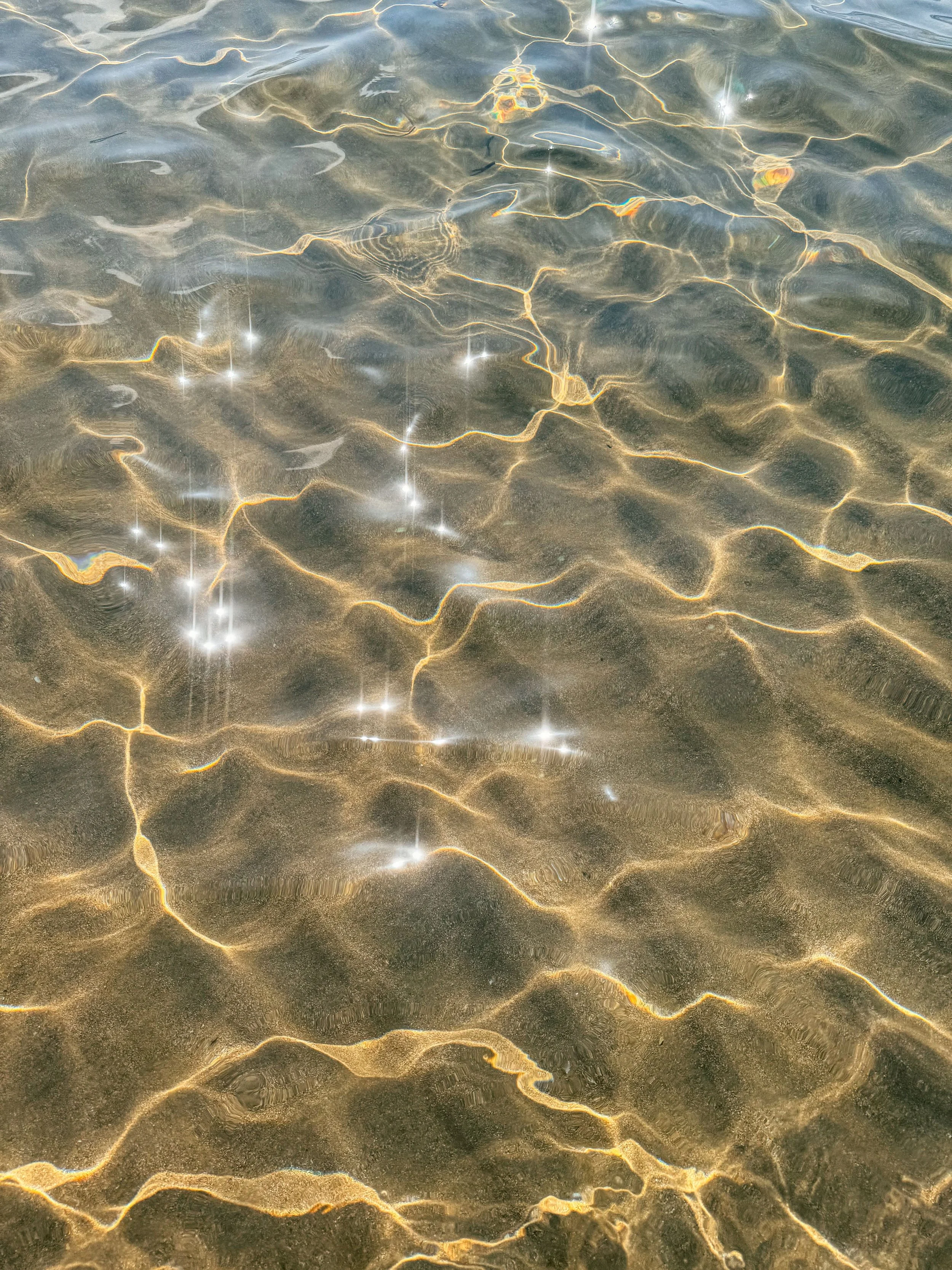 Close-up of shallow water with sunlight reflections, showing sandy bottom and light ripples on the surface.