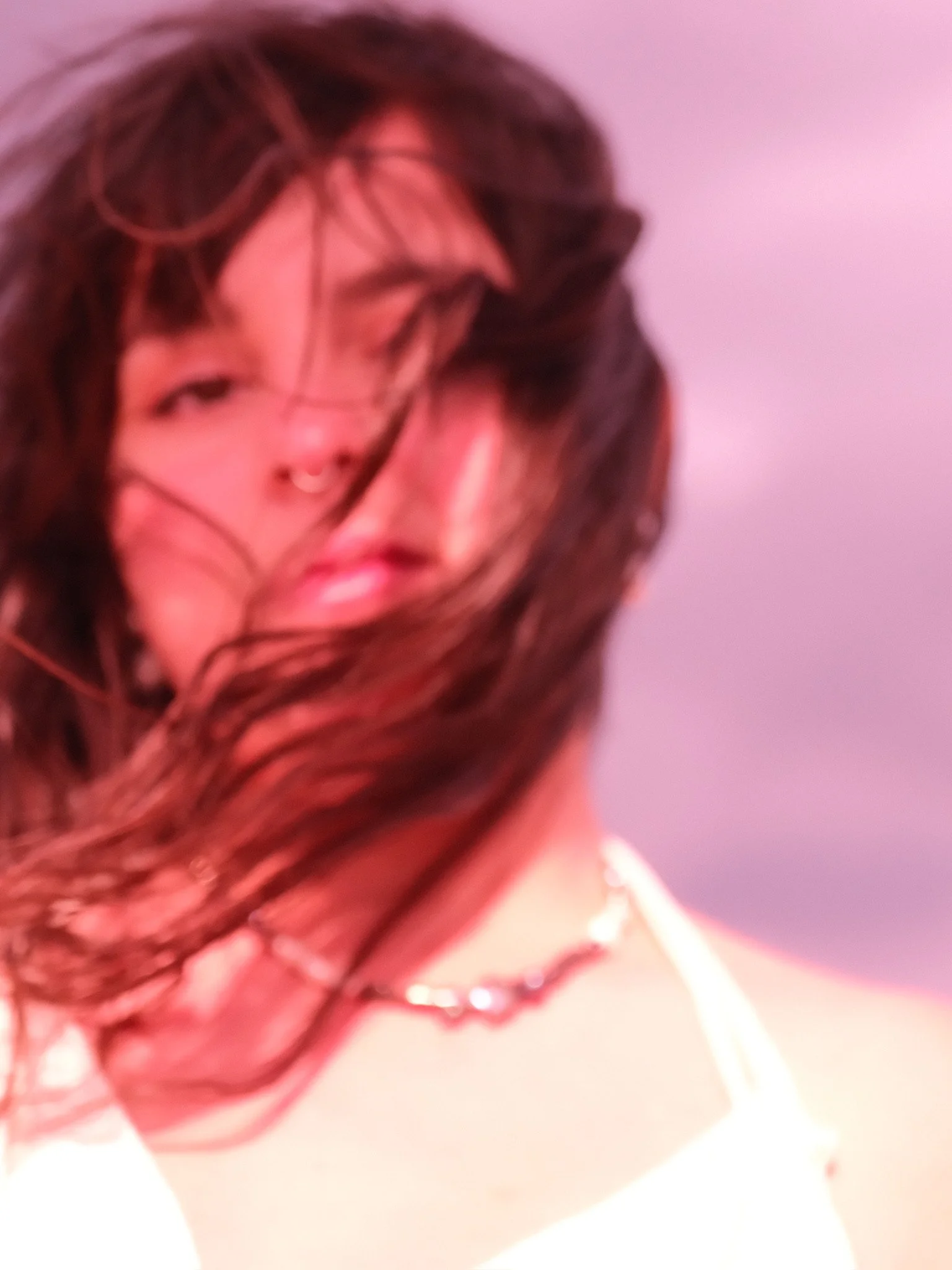 Close-up of a person's face with long dark hair and a nose ring, blurred with pink and purple lighting.