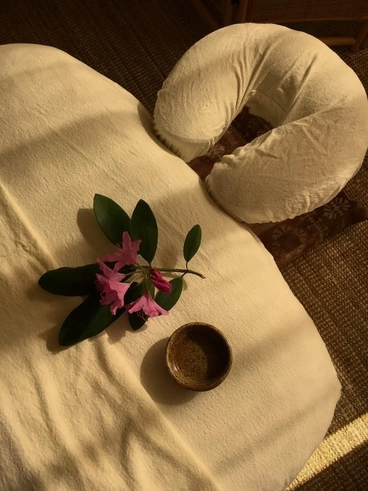 A plush U-shaped pillow, pink flowers with green leaves, and a small bowl on a cream-colored bedspread.