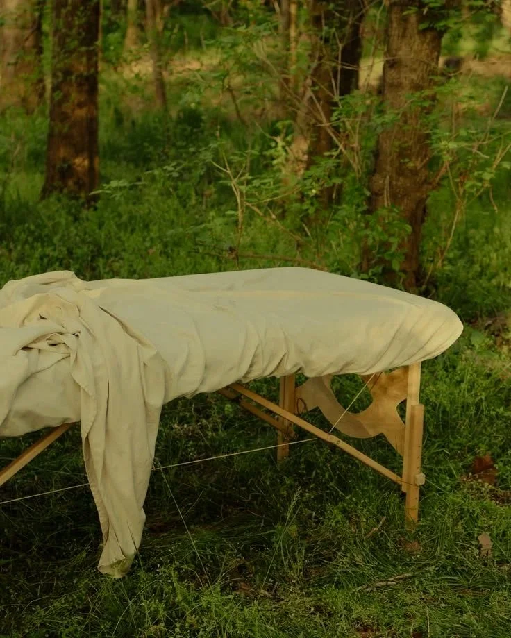 An outdoor scene in a forest with a wooden structure supporting a rolled-up mattress or padding draped with a beige cloth.
