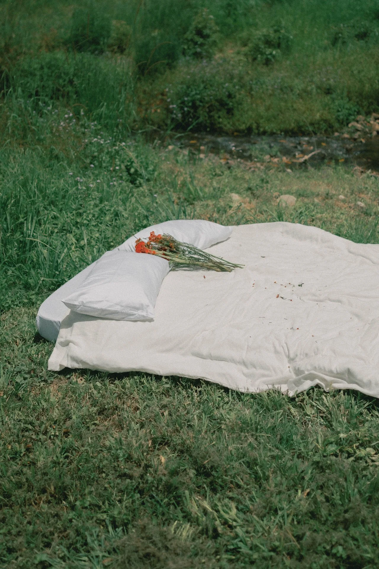 A white mattress with pillows and a bunch of orange flowers on top is placed outdoors on grass, near a small stream with green bushes in the background.