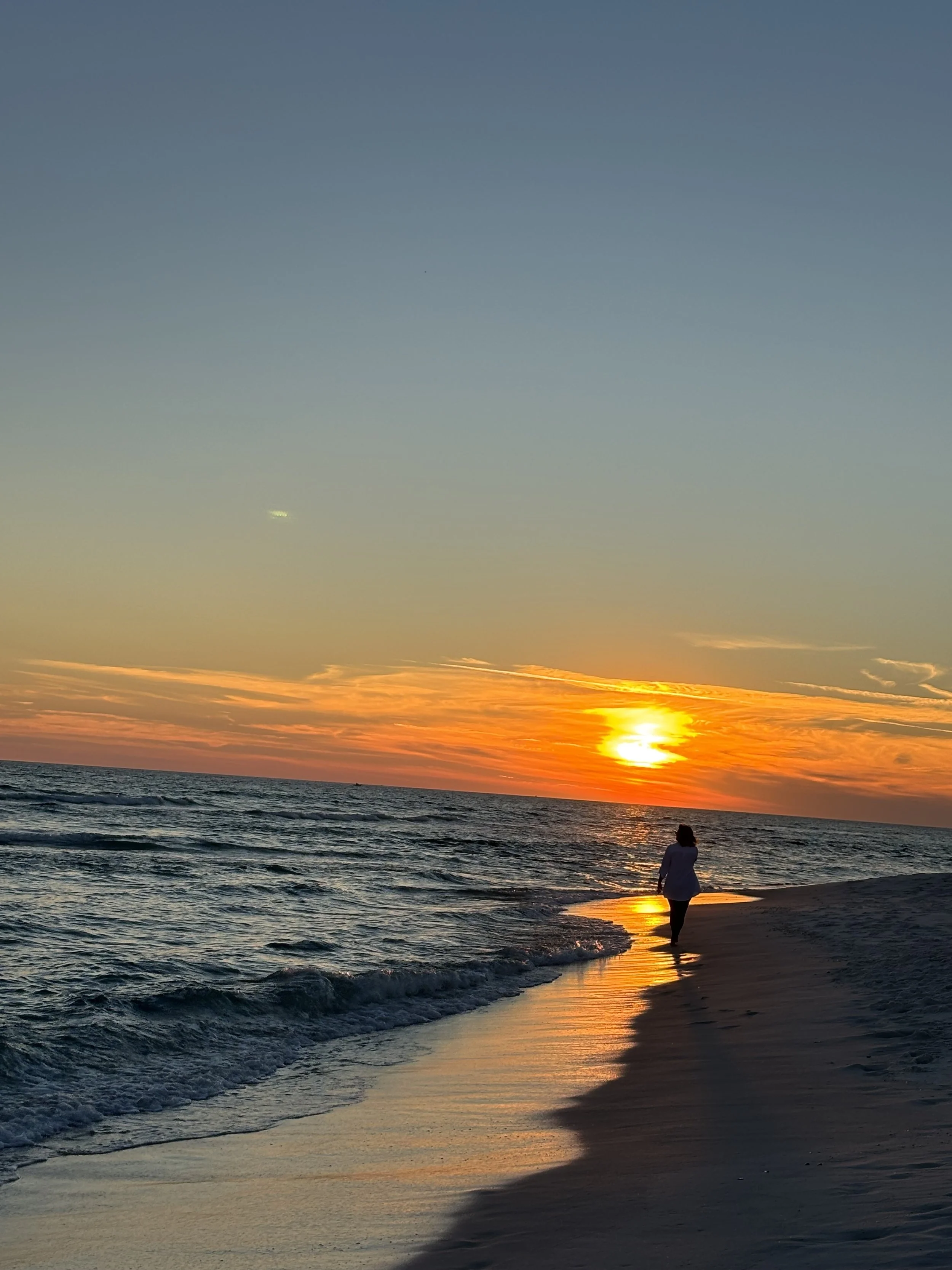A person walking along the beach during sunset with the sun near the horizon, casting orange and yellow hues in the sky and reflecting on the water.