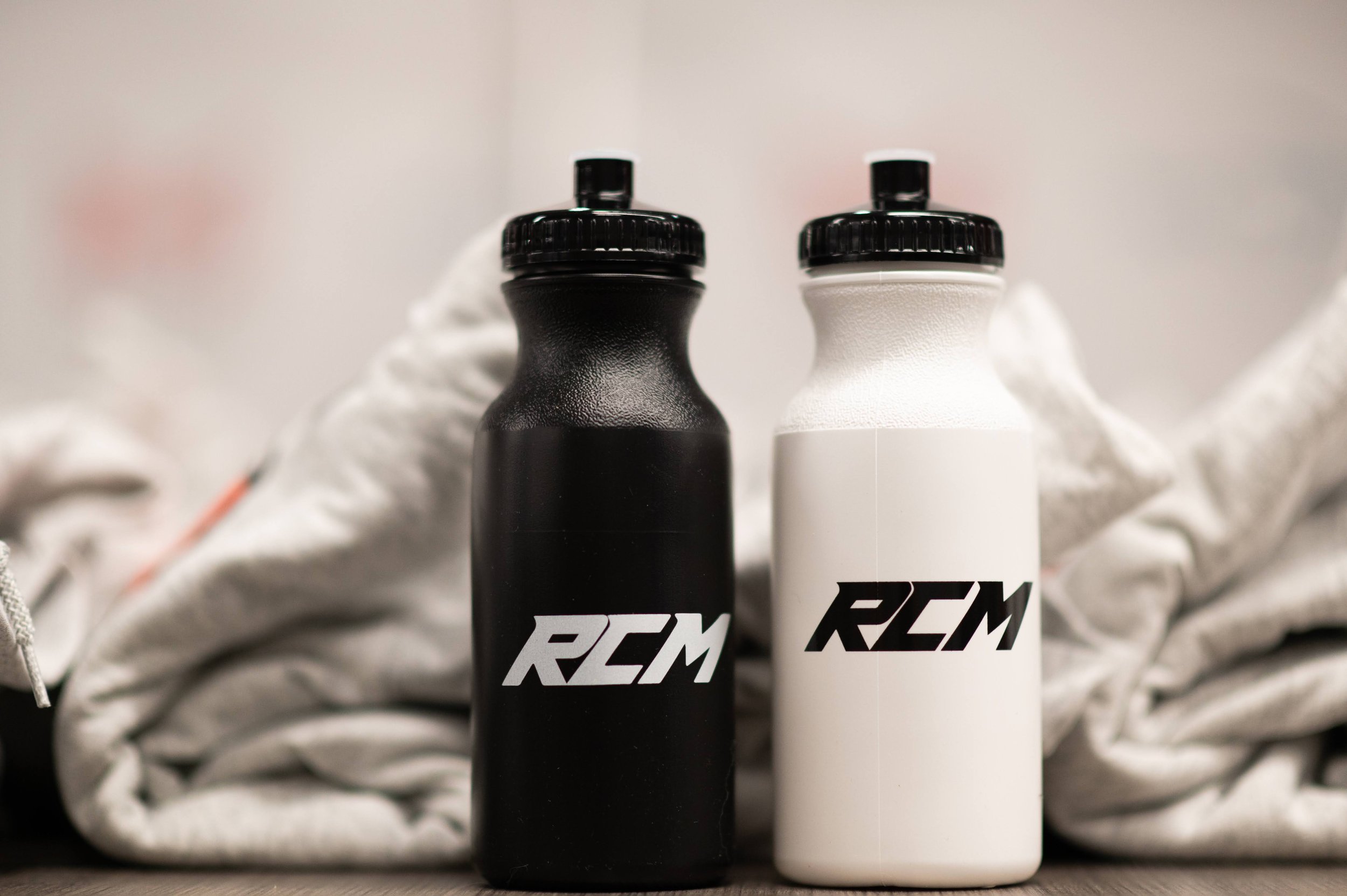 Two bottles, one black and one white, labeled with the RCM logo, positioned in front of a crumpled paper background.