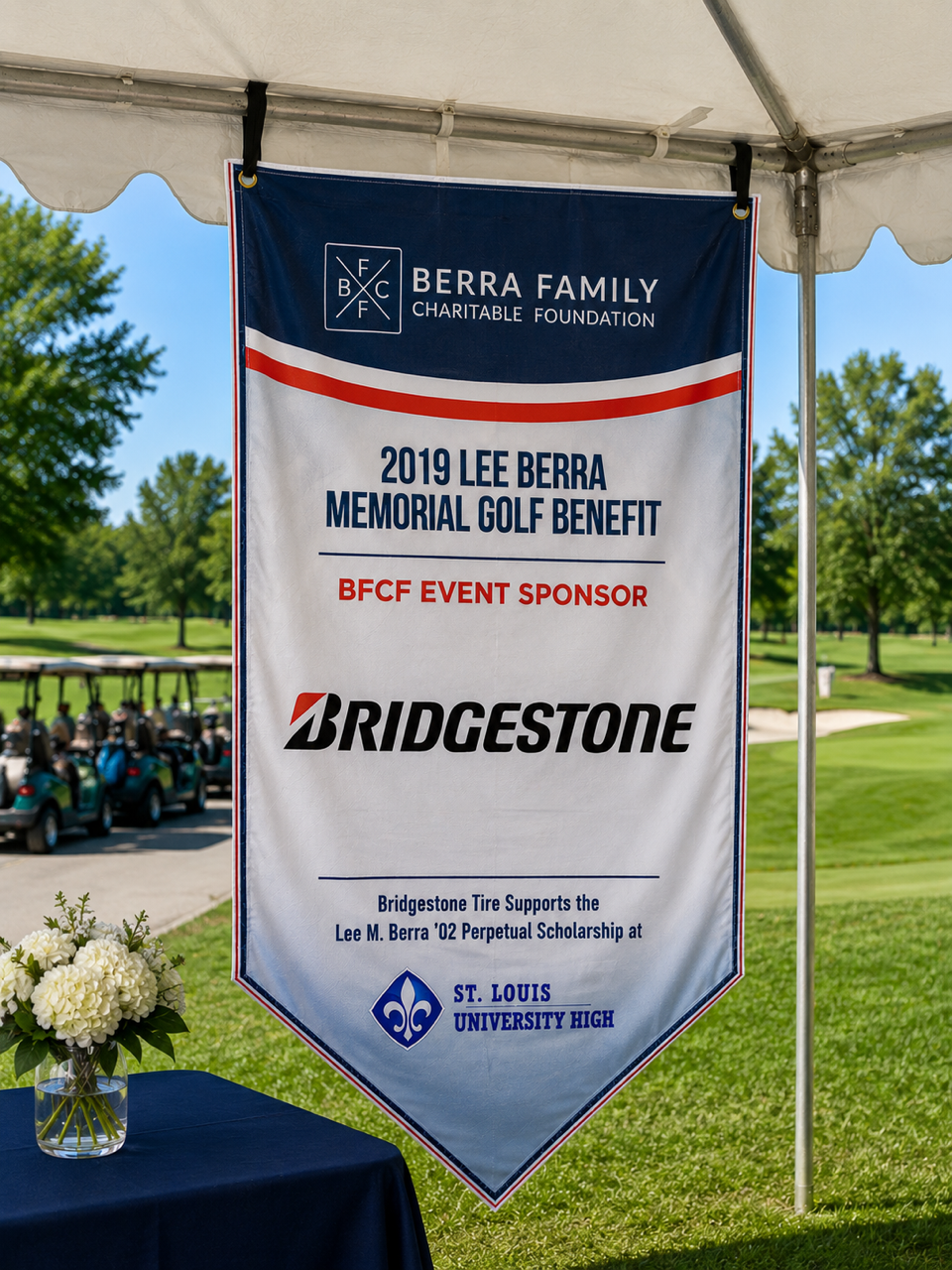 A banner hanging under a white tent at a golf course, displaying information about the 2019 Lee Berra Memorial Golf Benefit, sponsored by Bridgestone, supporting the Lee M. Berra '02 Perpetual Scholarship at St. Louis University High, with a table of