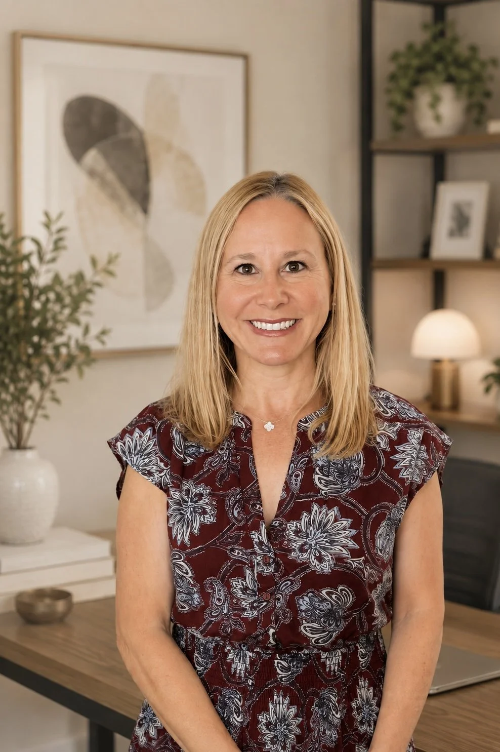A smiling woman with blonde hair wearing a maroon floral dress, standing in a modern, decorated room with framed artwork, a potted plant, and a lit table lamp.