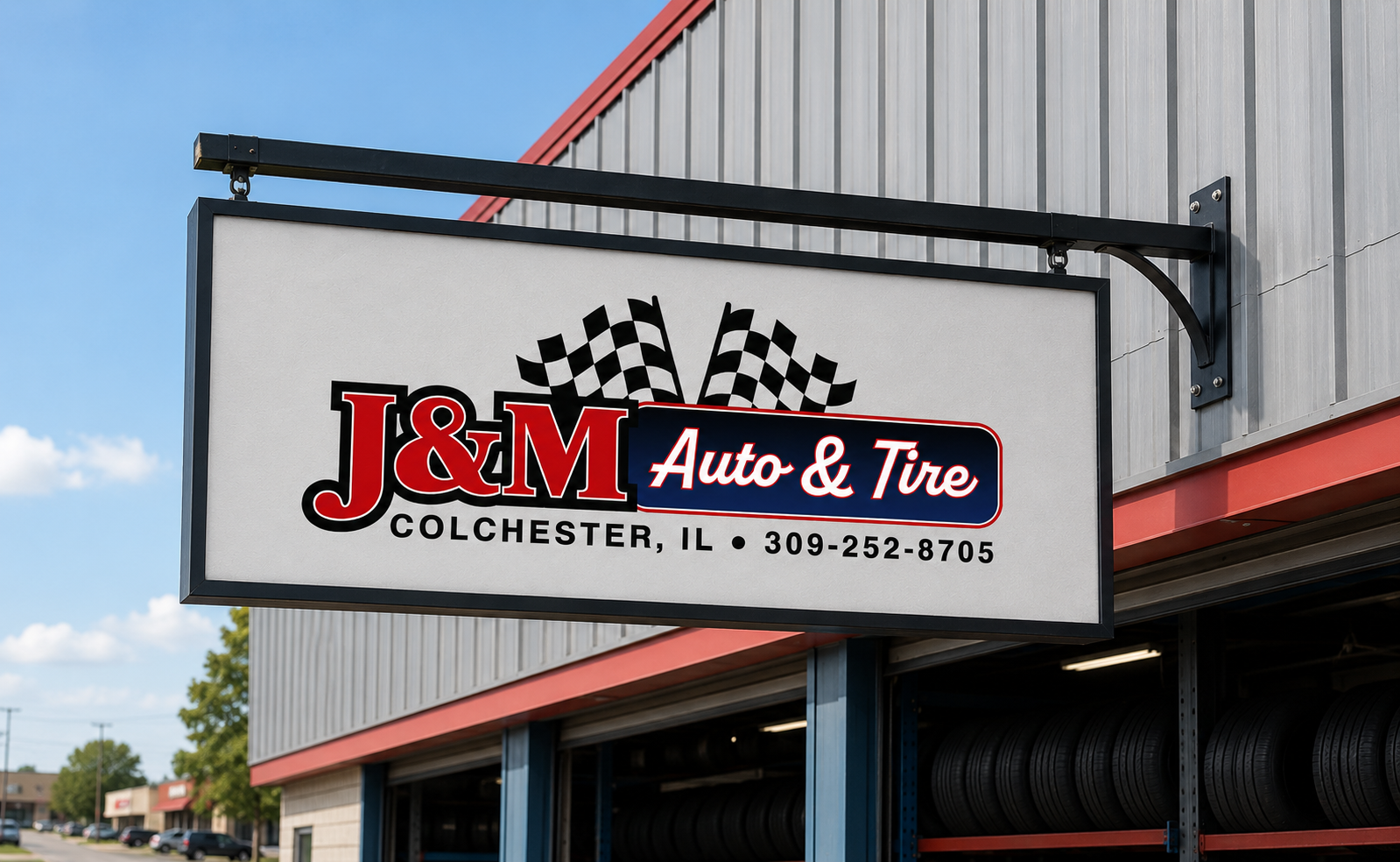 Sign for J&M Auto & Tire with checkered flags, located in Colchester, Illinois, with phone number 309-252-8705, outside of a building with tires visible underneath.