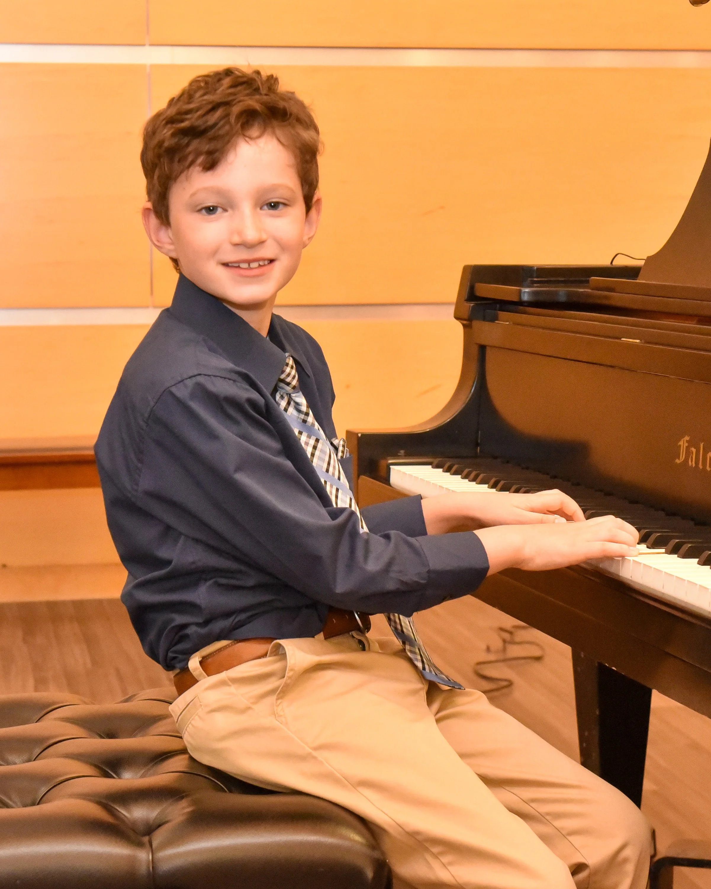 A young boy with short curly brown hair playing a black grand piano in a room with wooden panel walls. He is smiling, wearing a dark blue shirt, khaki pants, and a striped tie, sitting on a brown plush tufted bench.