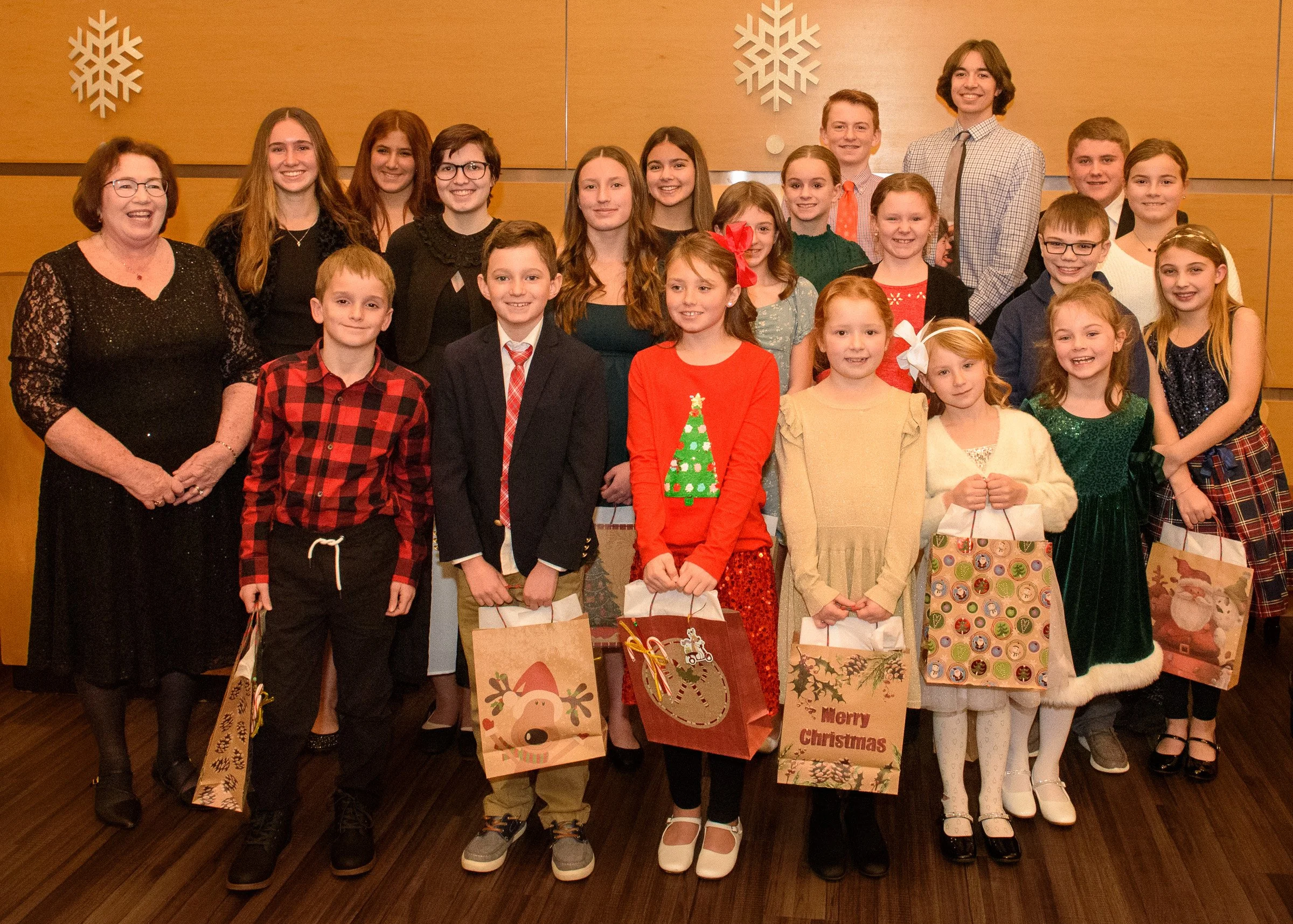 Group of children and adults celebrating Christmas indoors, holding holiday gift bags, with Christmas-themed decorations on the wall.