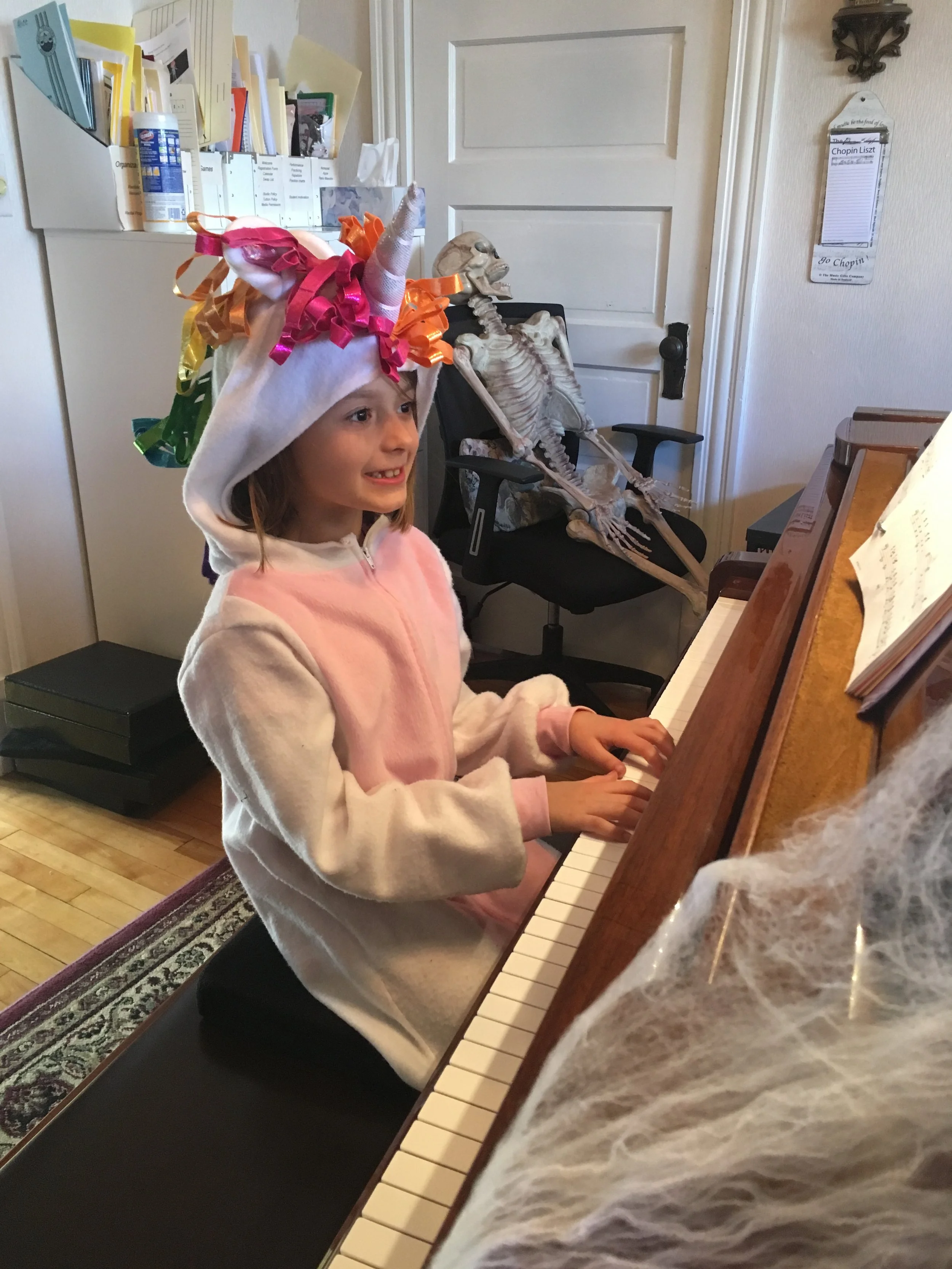 A young girl wearing a unicorn headband decorated with colorful ribbons and a pink fleece jacket, smiling while playing the piano. A skeleton decoration is seated in a chair behind her in the background.