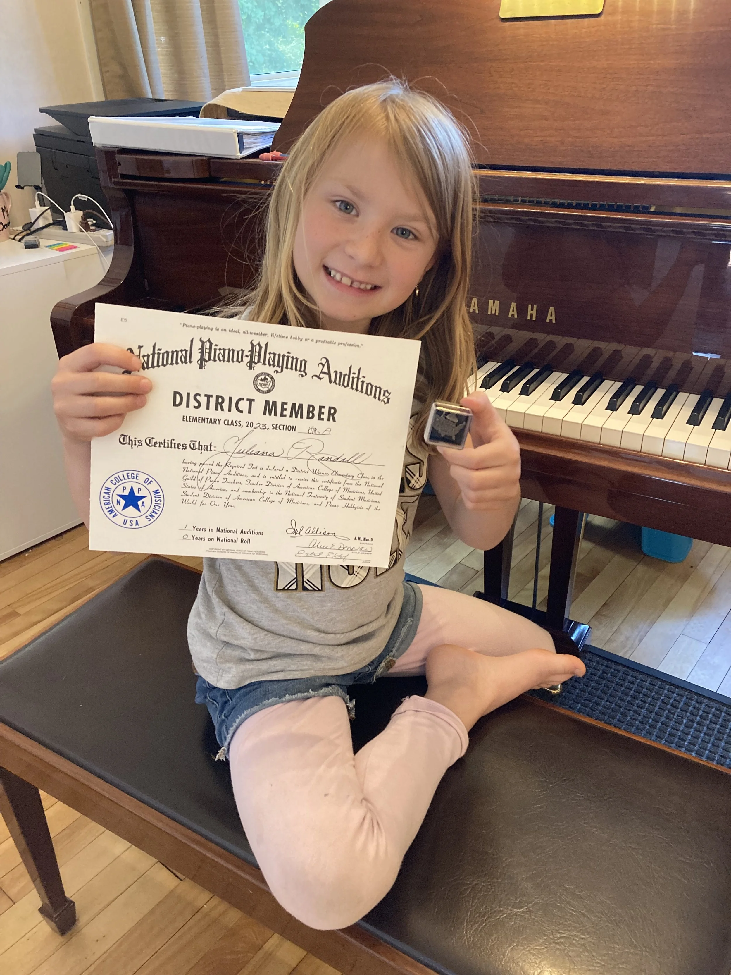 A young girl sitting on a drafting table with a Yamaha piano in the background, holding a certificate and a small award, smiling at the camera.