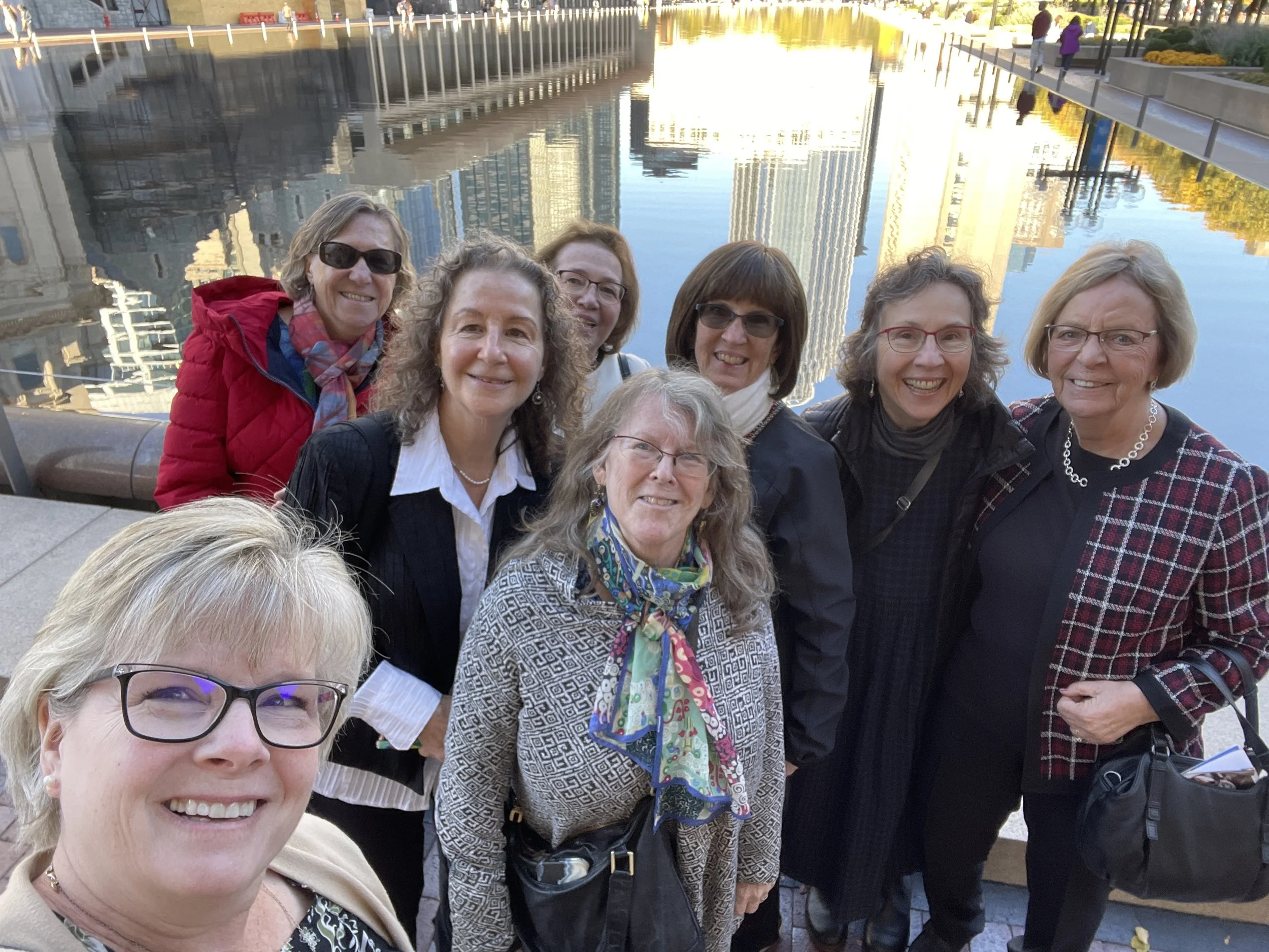 A group of women taking a selfie outdoors near a water feature with tall buildings and trees in the background.