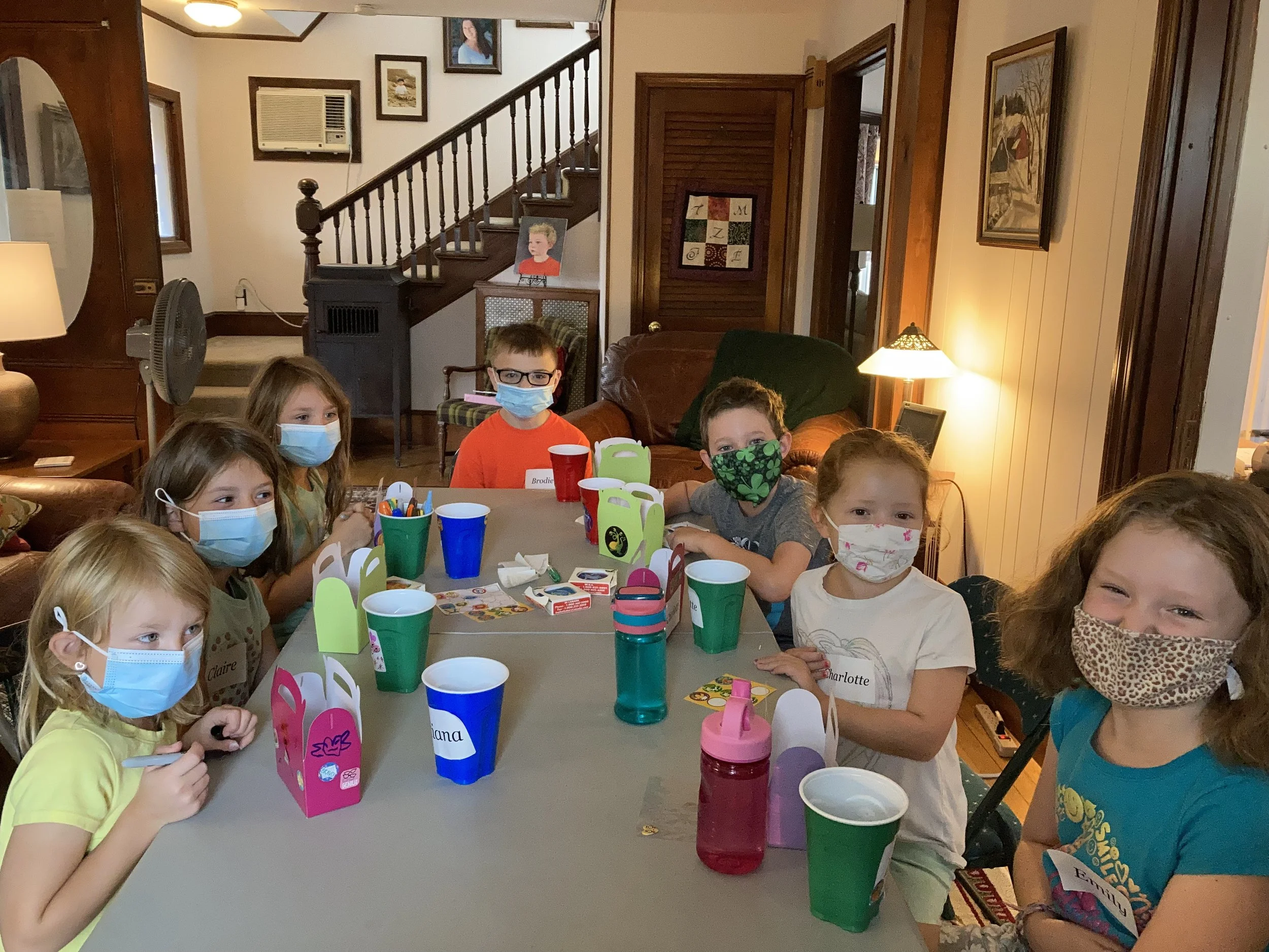 Children sitting around a table with colorful cups and party favors, all wearing masks, in a cozy living room with wood-paneled walls and framed artwork.