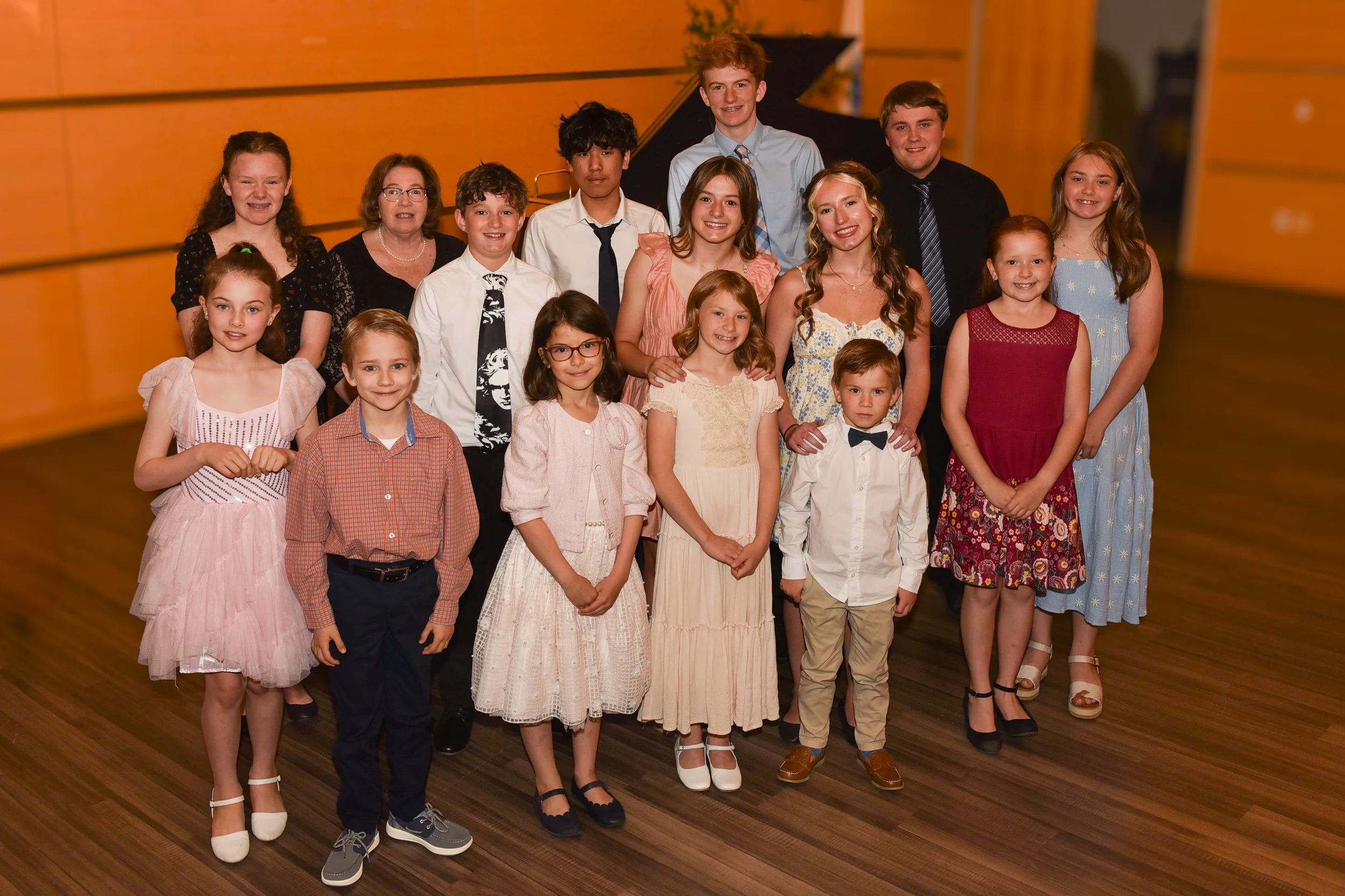 A group of 14 children and teenagers, mostly girls, dressed in semi-formal attire, standing in a room with wooden walls and floor, and a black grand piano in the background. They are smiling and posing for the picture.