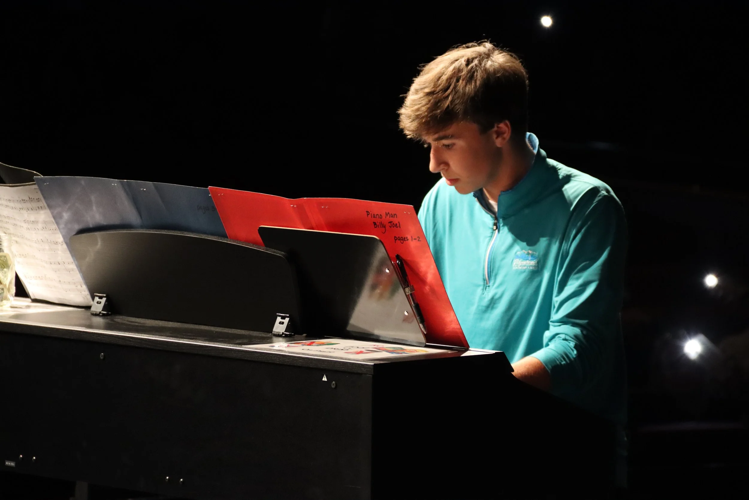 Young man playing piano in a dark setting, focused on sheet music labeled 'Piano Man Billy Joel pages 1-2'.