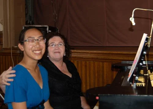 Two women sitting at a piano in a room with wooden paneling, smiling at the camera.