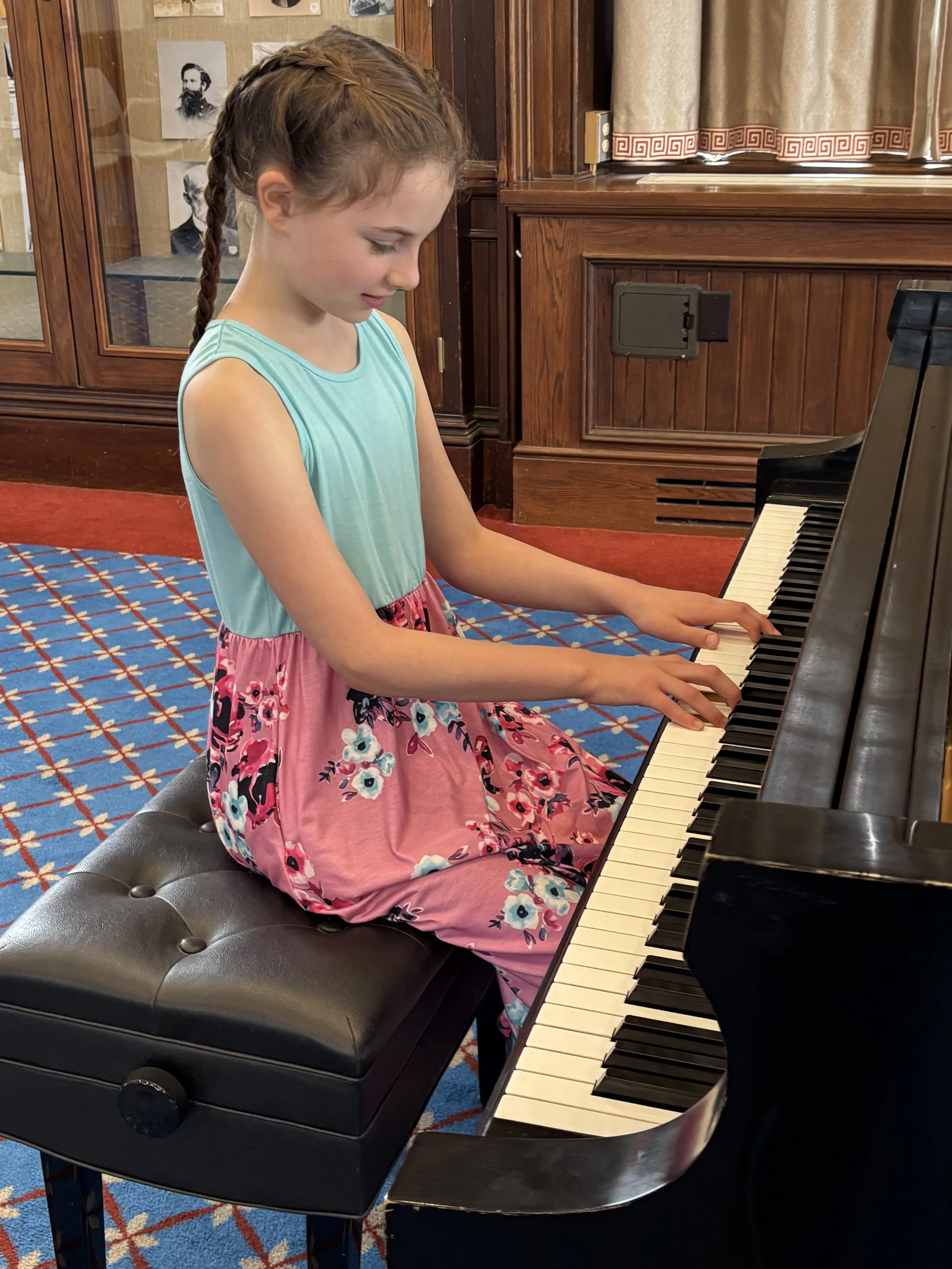 A young girl with braided brown hair wearing a turquoise top and a pink floral skirt playing a black piano in a room with wooden paneling and pictures on the wall.