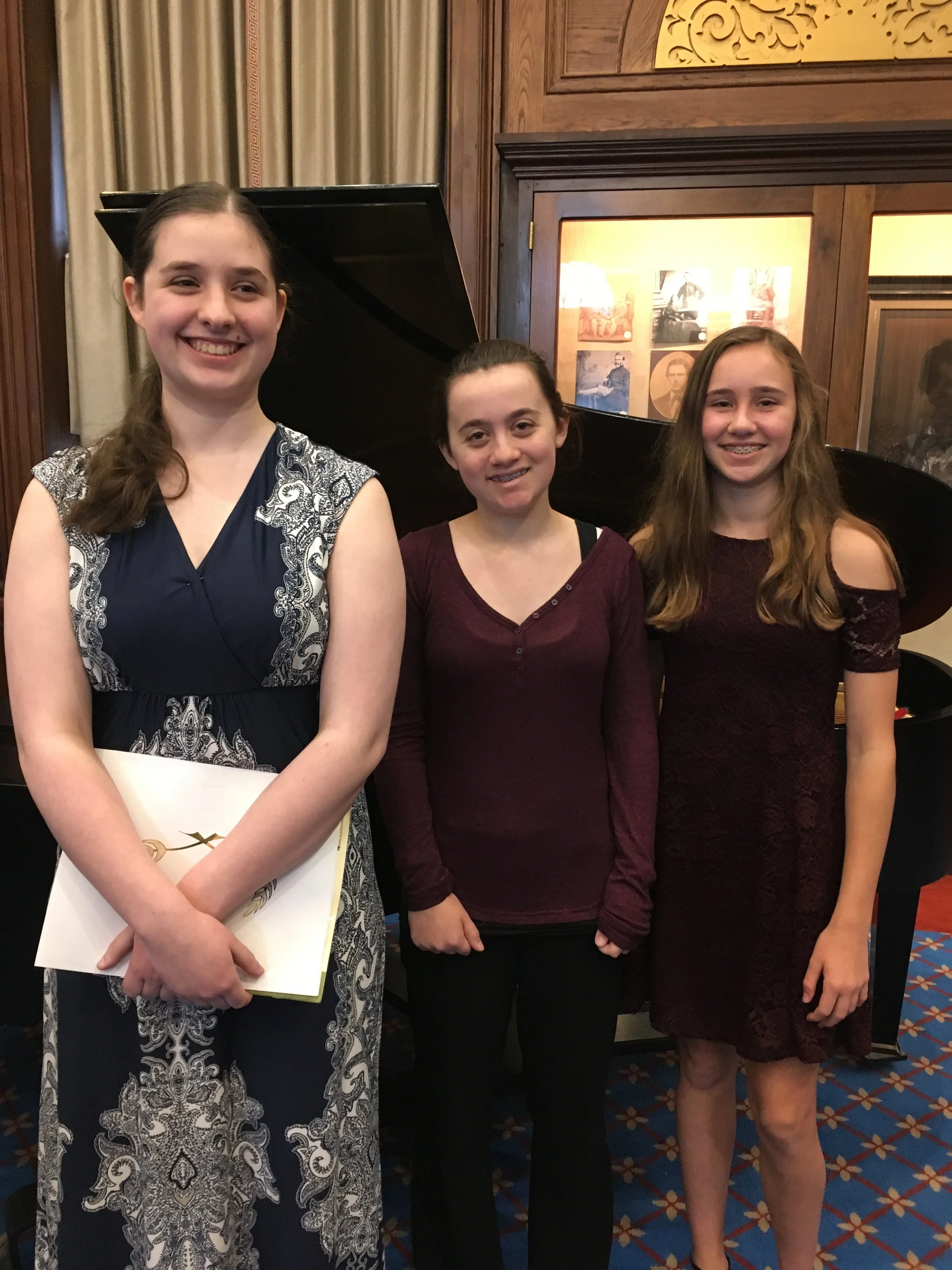 Three young girls standing indoors in front of a piano, smiling at the camera. The girl on the left is wearing a dress with black and white pattern, holding some papers. The girl in the middle has dark hair tied back, wearing a maroon top, and the girl on the right has wavy light brown hair, wearing a maroon dress.