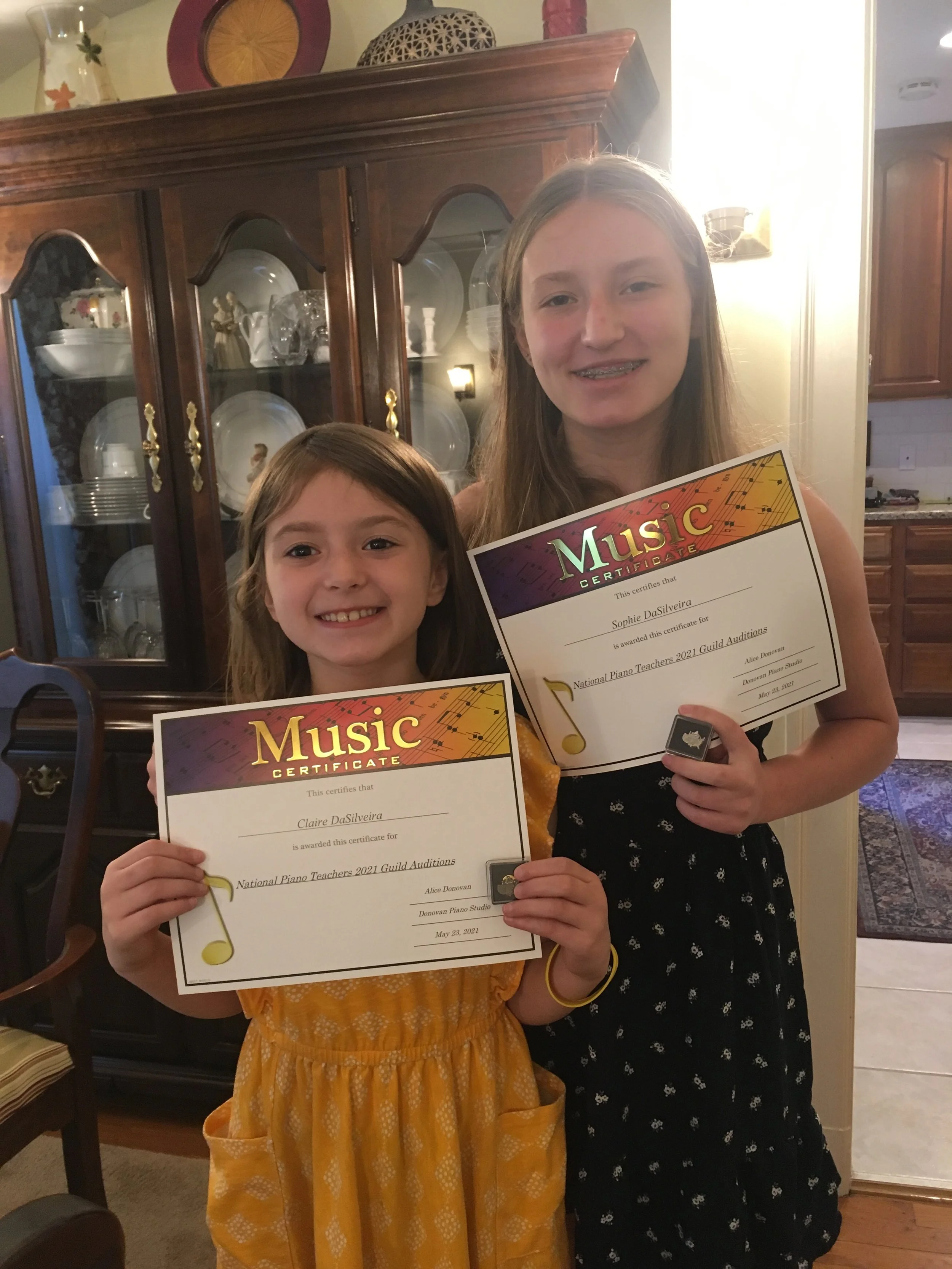 Two young girls standing indoors, holding certificates for a music audition. They are smiling, with a china cabinet filled with dishes and decorative items behind them, in a warmly lit room.