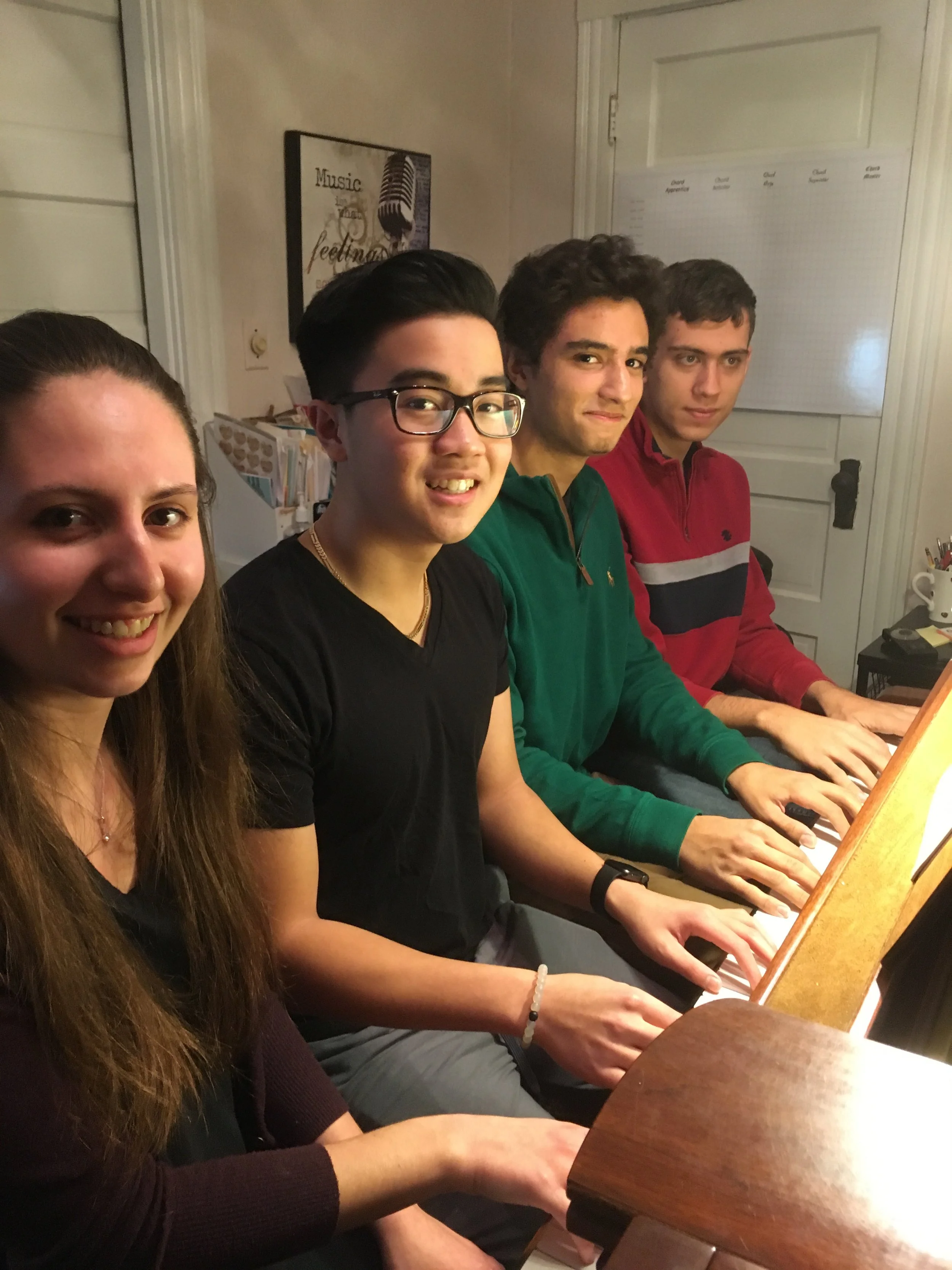 Four young people sitting at a piano, all appearing to play or listen to music, in a cozy indoor setting.