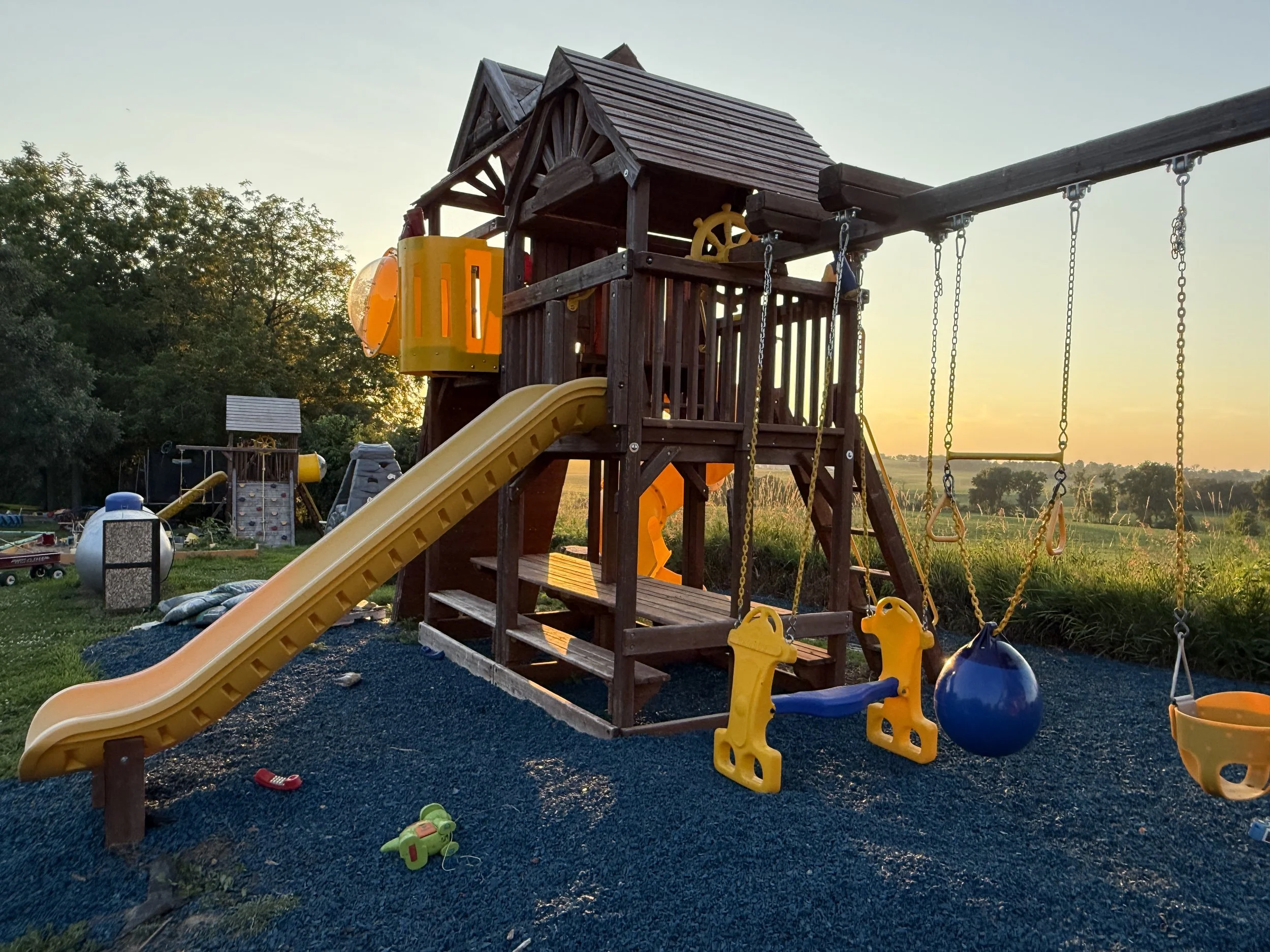 A backyard wooden playground set with a slide, swings, and a climbing wall, illuminated by sunset light.