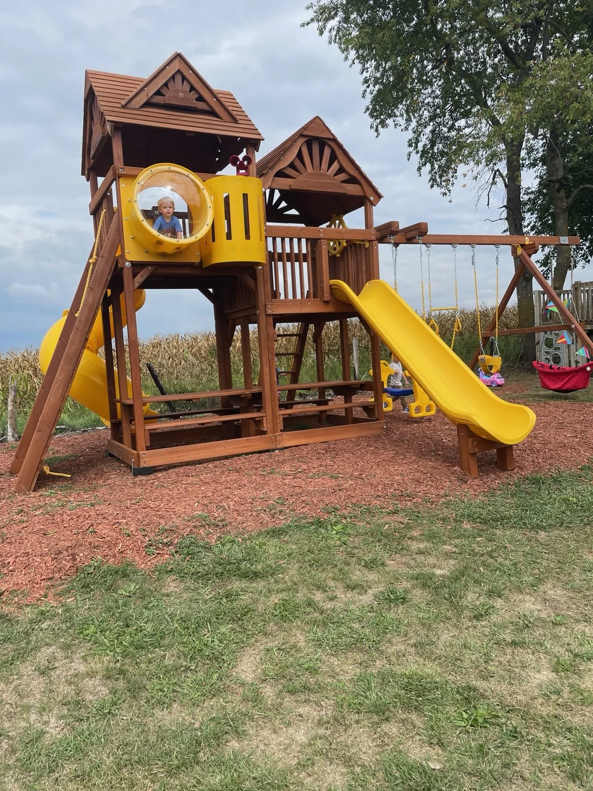 A children's wooden playset with a yellow slide, a yellow tunnel, swings, and a castle-like structure on a grassy and dirt area with trees and a cloudy sky in the background.