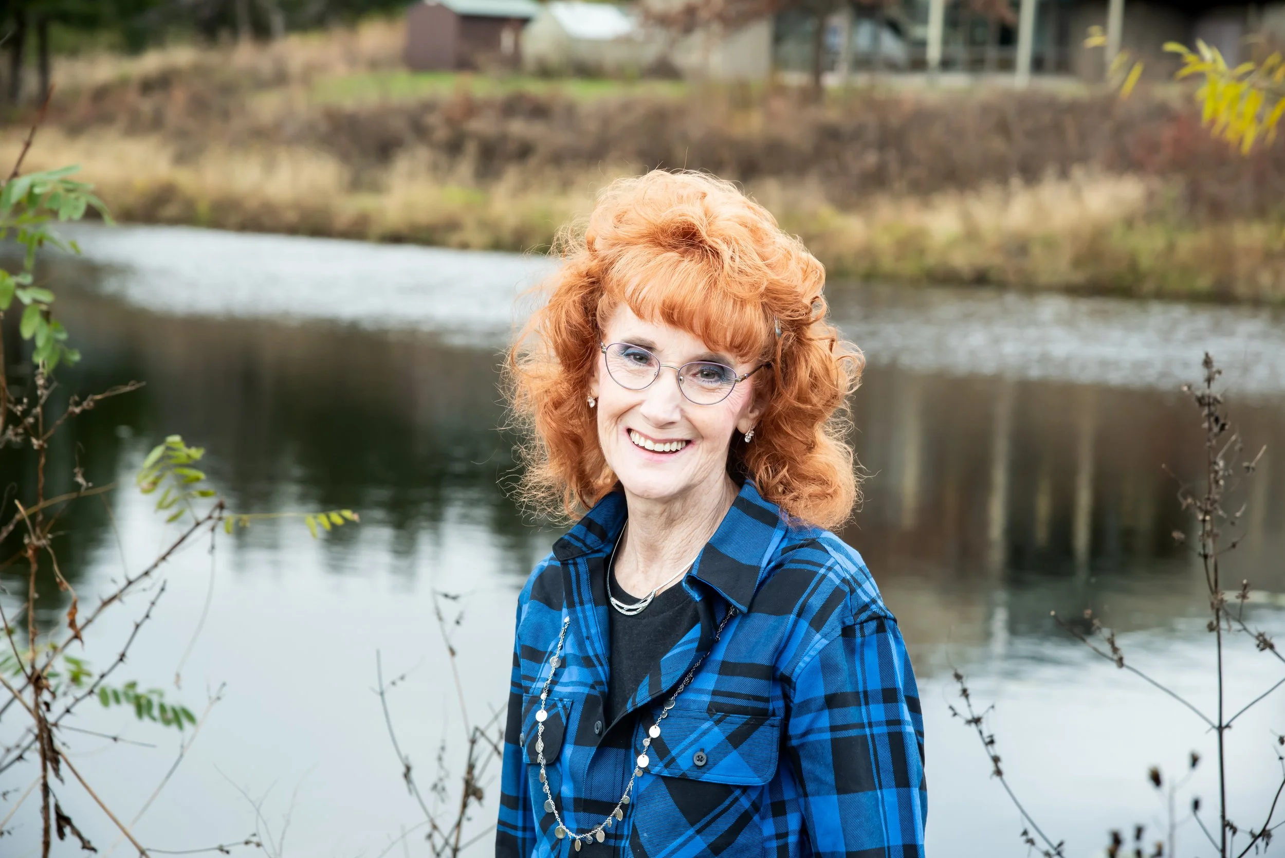 A woman with curly red hair, glasses, and a blue plaid shirt standing near a river, smiling at the camera with a background of water and trees.
