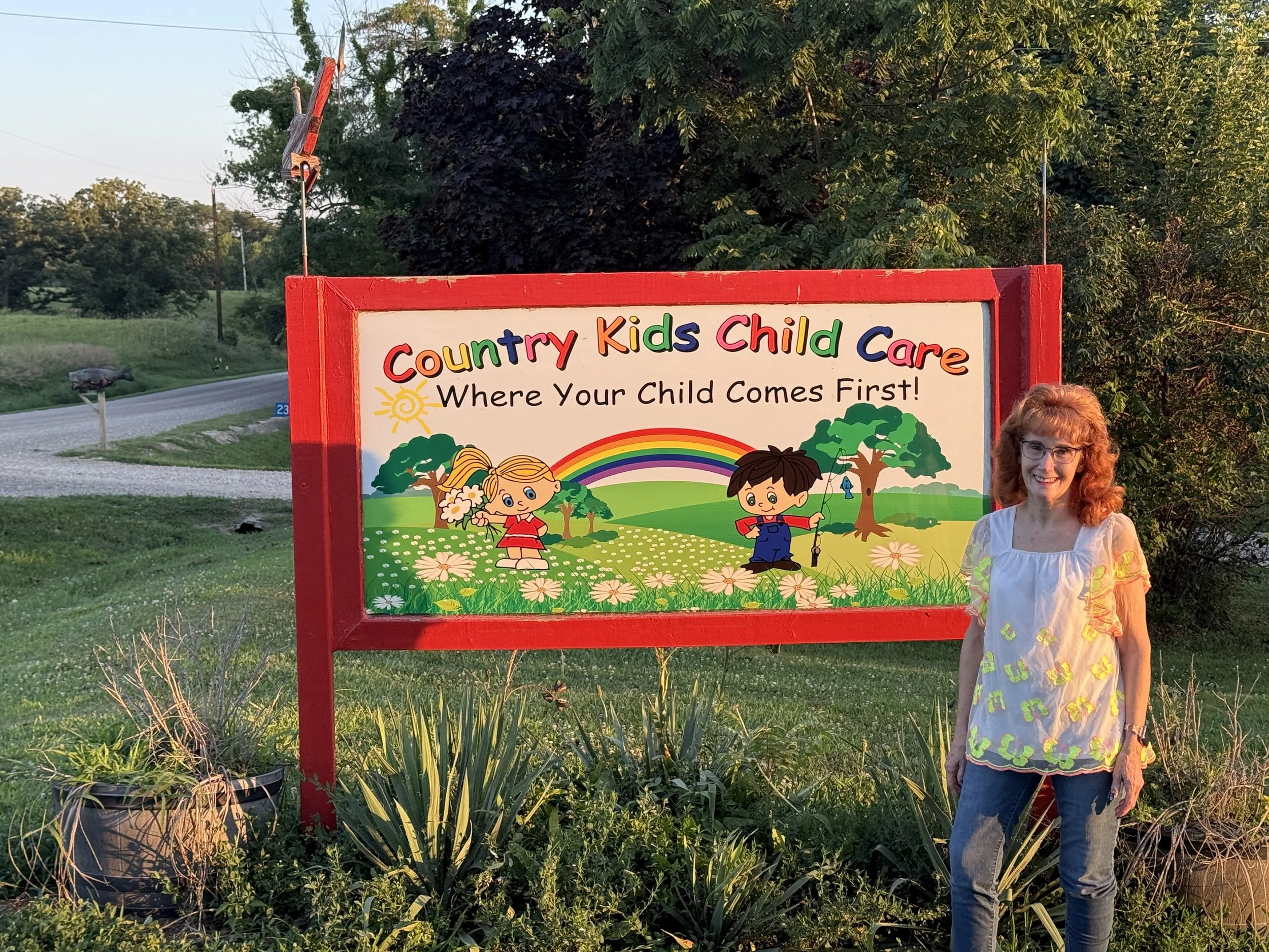 A woman with red hair and glasses standing next to a colorful sign for a children's daycare called 'Country Kids Child Care'. The sign features cartoon children, trees, a rainbow, and a sun, with the slogan 'Where Your Child Comes First!'. The woman is smiling and wearing a white top with colorful patterns. The setting is outdoors with green grass, plants, and trees in the background, during what appears to be late afternoon or early evening.