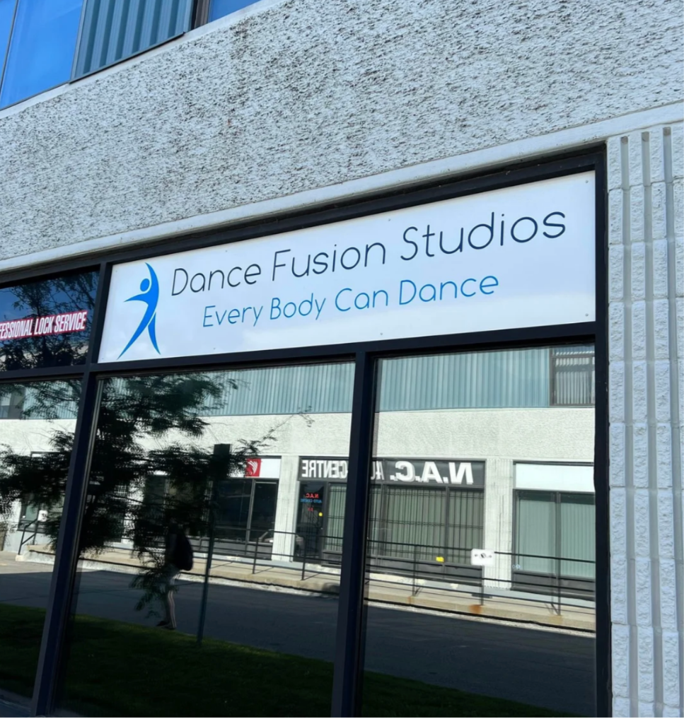 Sign for Dance Fusion Studios with the tagline "Every Body Can Dance" on a building exterior, reflecting street and building in the glass windows.
