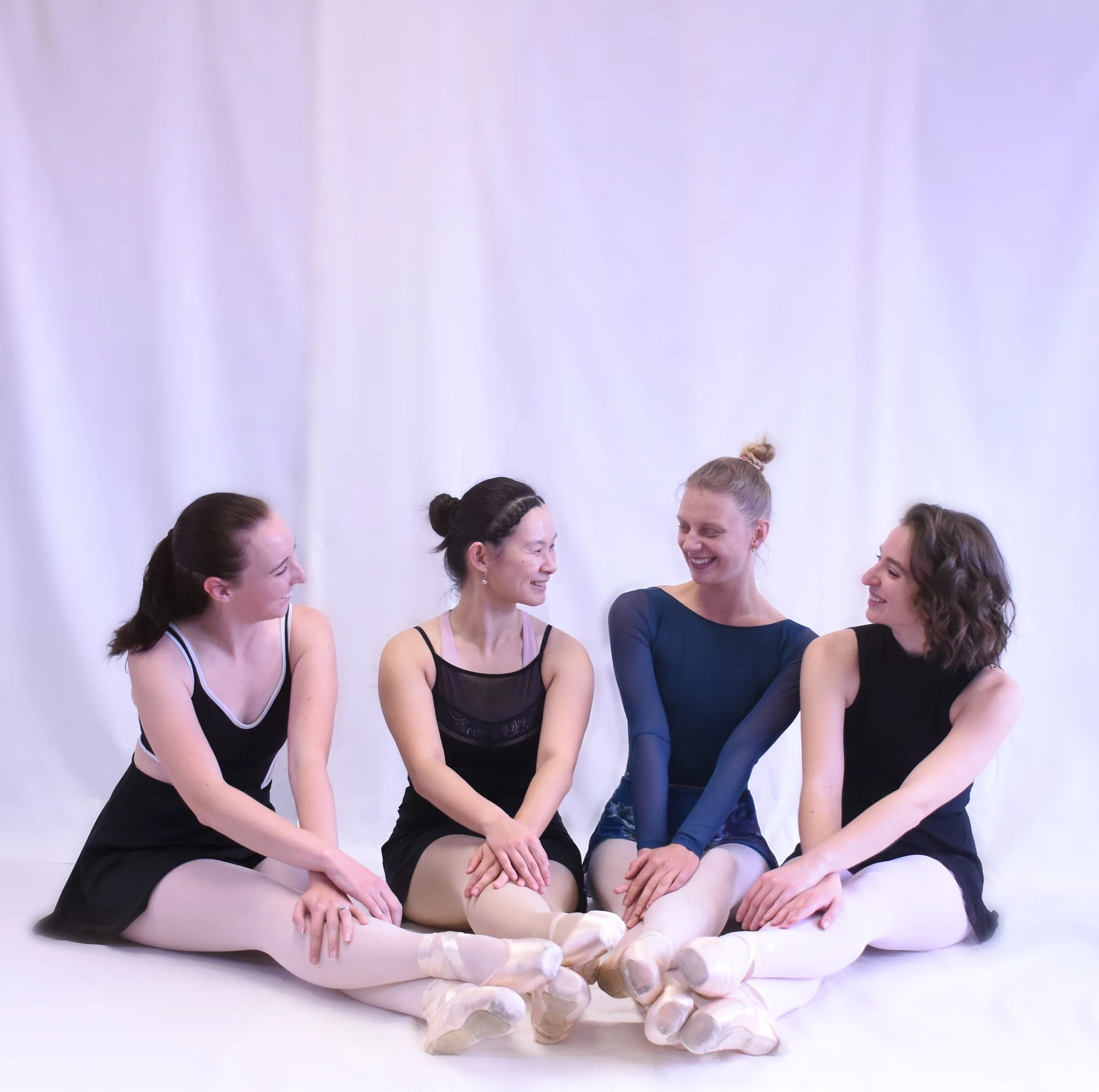A group of ballet dancers in dancewear, sitting together and smiling in front of a plain white background.