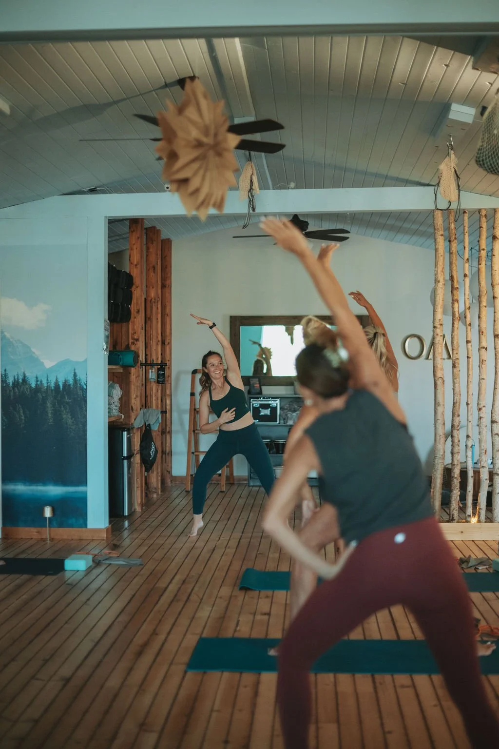 People practicing yoga in a cozy, wood-paneled indoor space with a mountain mural, ceiling fans, and natural decor.