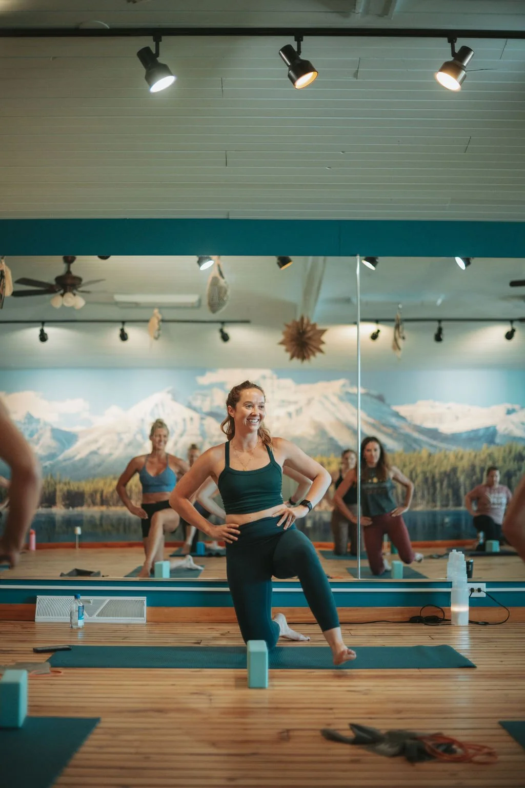 A woman leading a yoga class in a studio with a mountain landscape mural on the wall. She is smiling and standing on one leg with one hand on her hip and the other on her waist. Other women are practicing yoga behind her, all on mats.