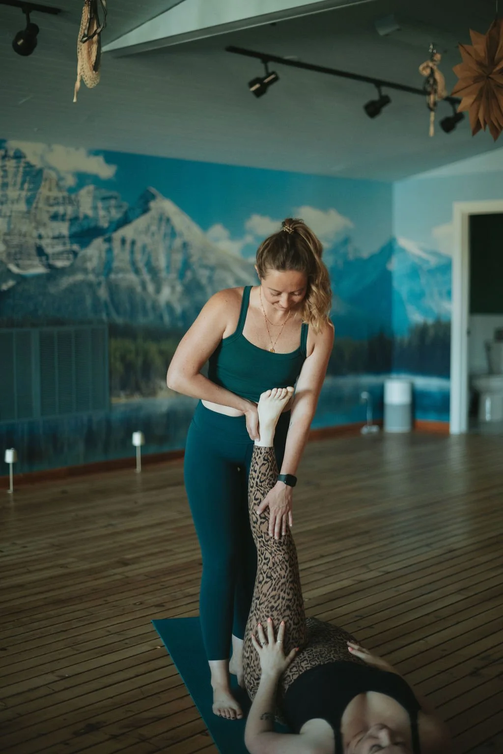 Yoga instructor assisting a woman in a yoga pose in a studio with mountain landscape mural